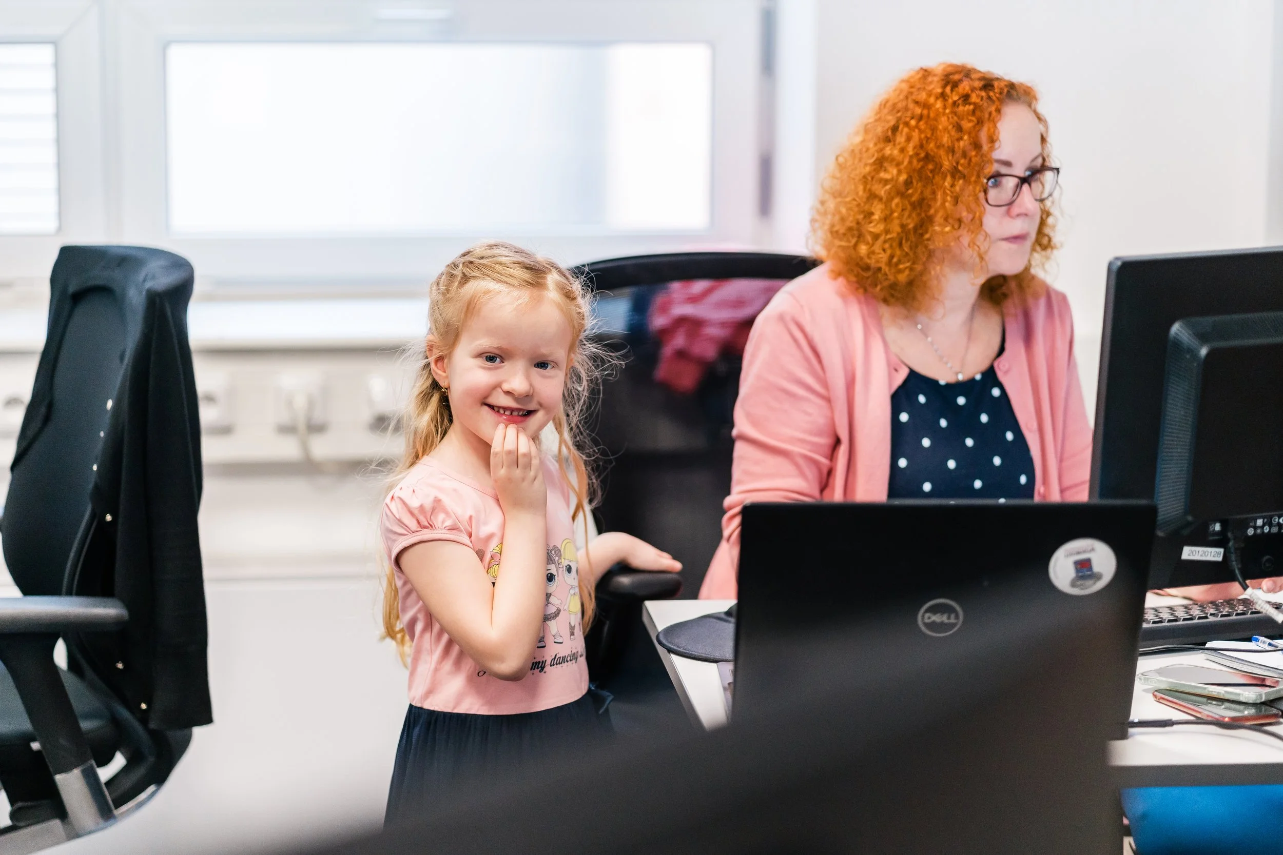 A young girl with long blonde hair smiling and touching her face, sitting at a desk next to a woman with curly red hair and glasses working on a computer in an office with large windows. Kids in the office in Switzerland.