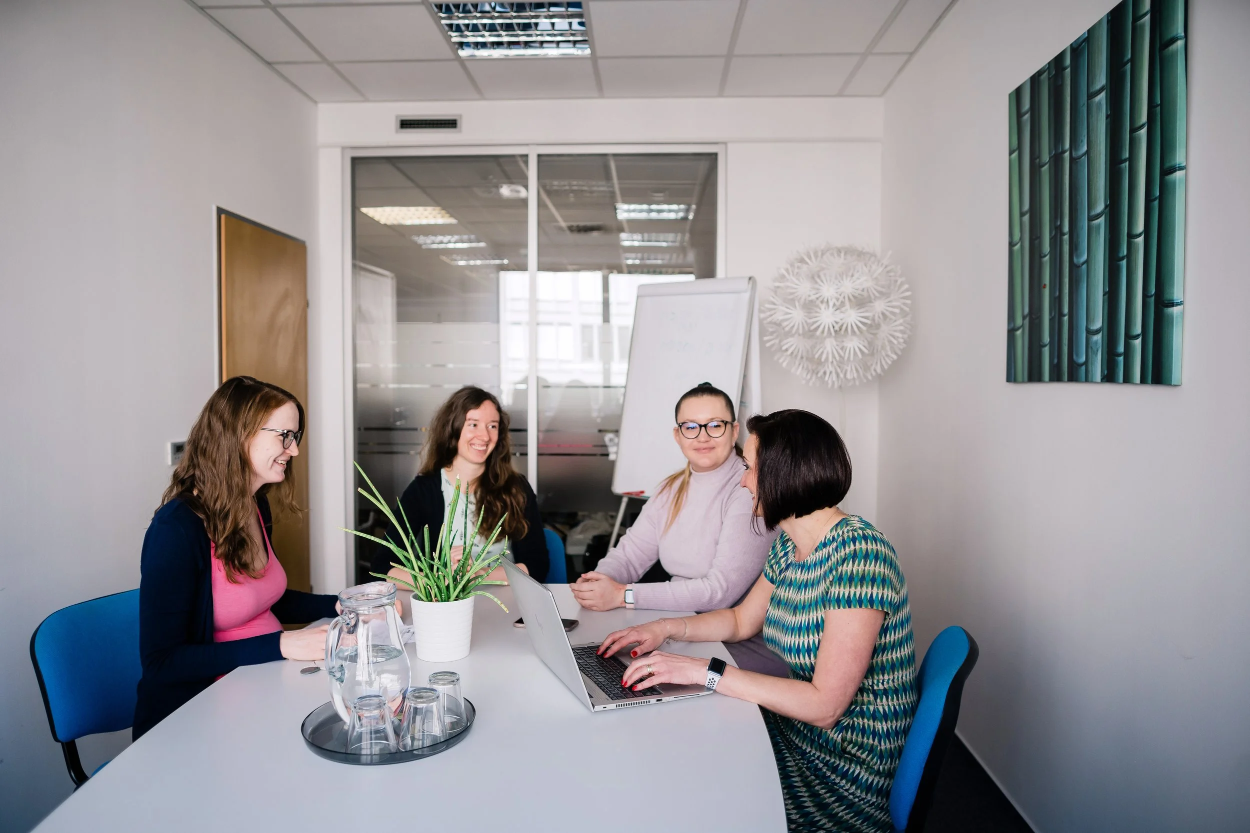 Four women sitting around a white conference table in a modern office, engaged in a discussion, with a laptop, a large plant, and glasses on the table. Employer branding photography in Zürich, Zug, Bern, Lucerne, Basel, St. Gallen - Switzerland.
