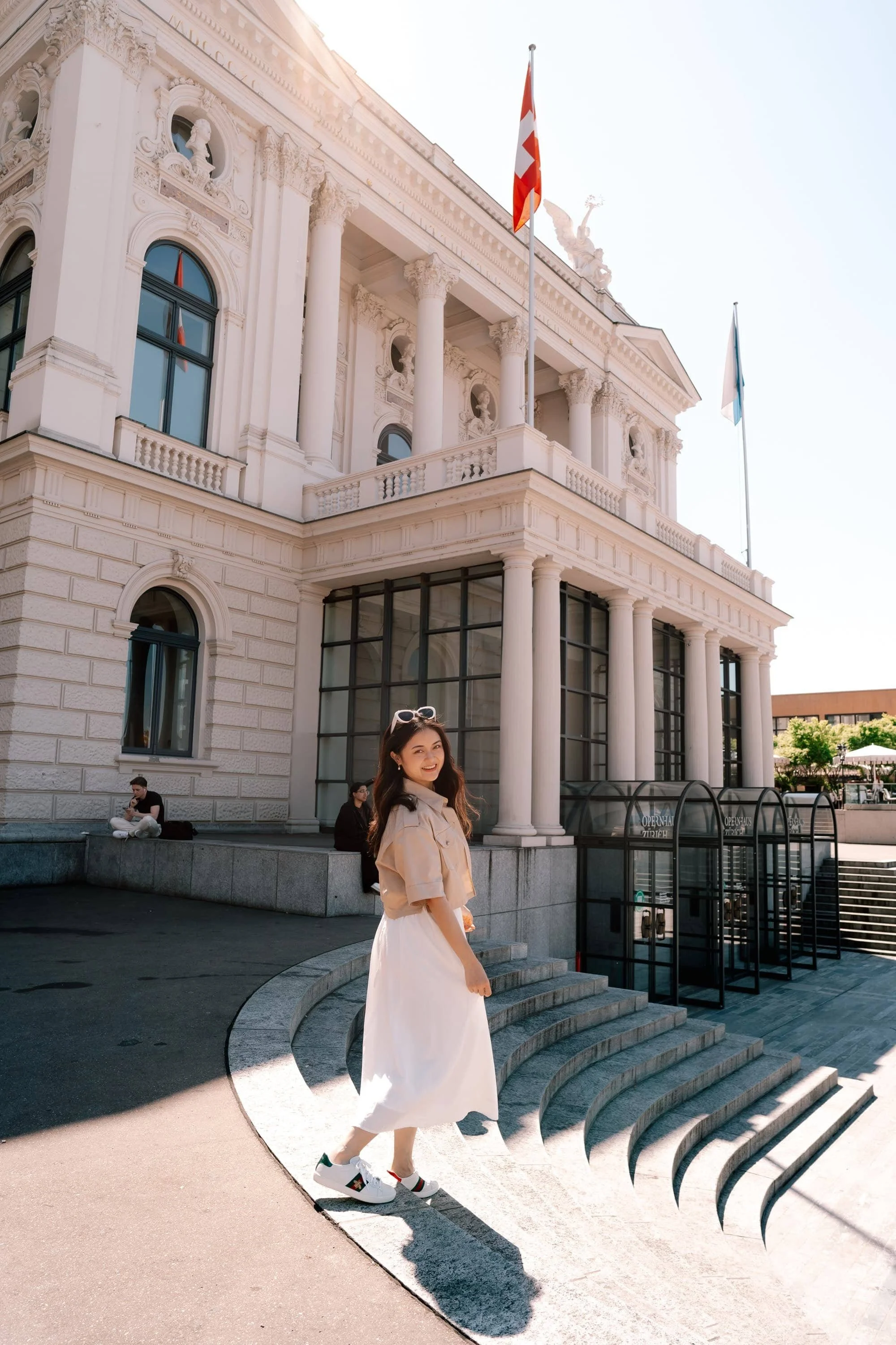 Woman in white skirt walking down the Opernhaus Zurich in Switzerland, smiling to the camera, posing for photoshoot in beautiful sunlight.