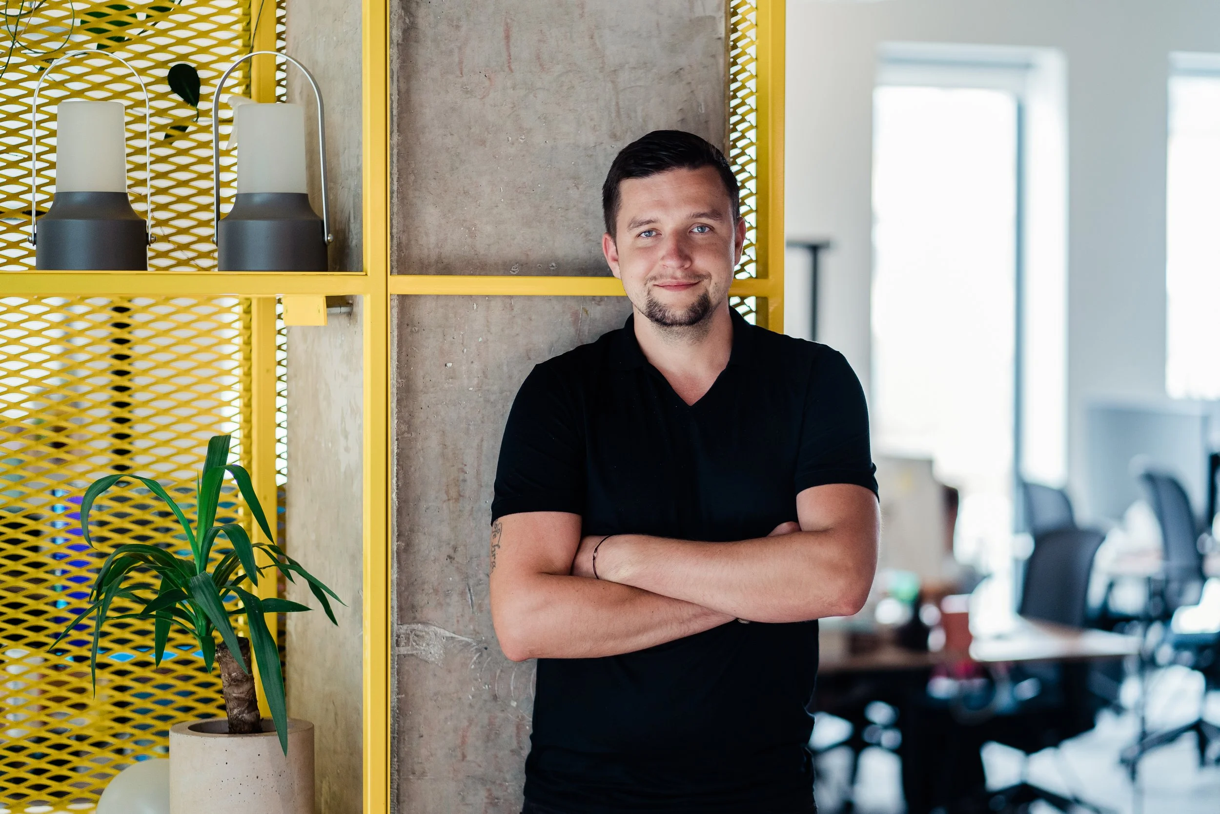 A young man with crossed arms and a slight smile standing in a modern office space. He has short dark hair, a beard, and is wearing a black shirt. There are yellow shelves with two lamps and a potted plant, and office desks visible in the background.