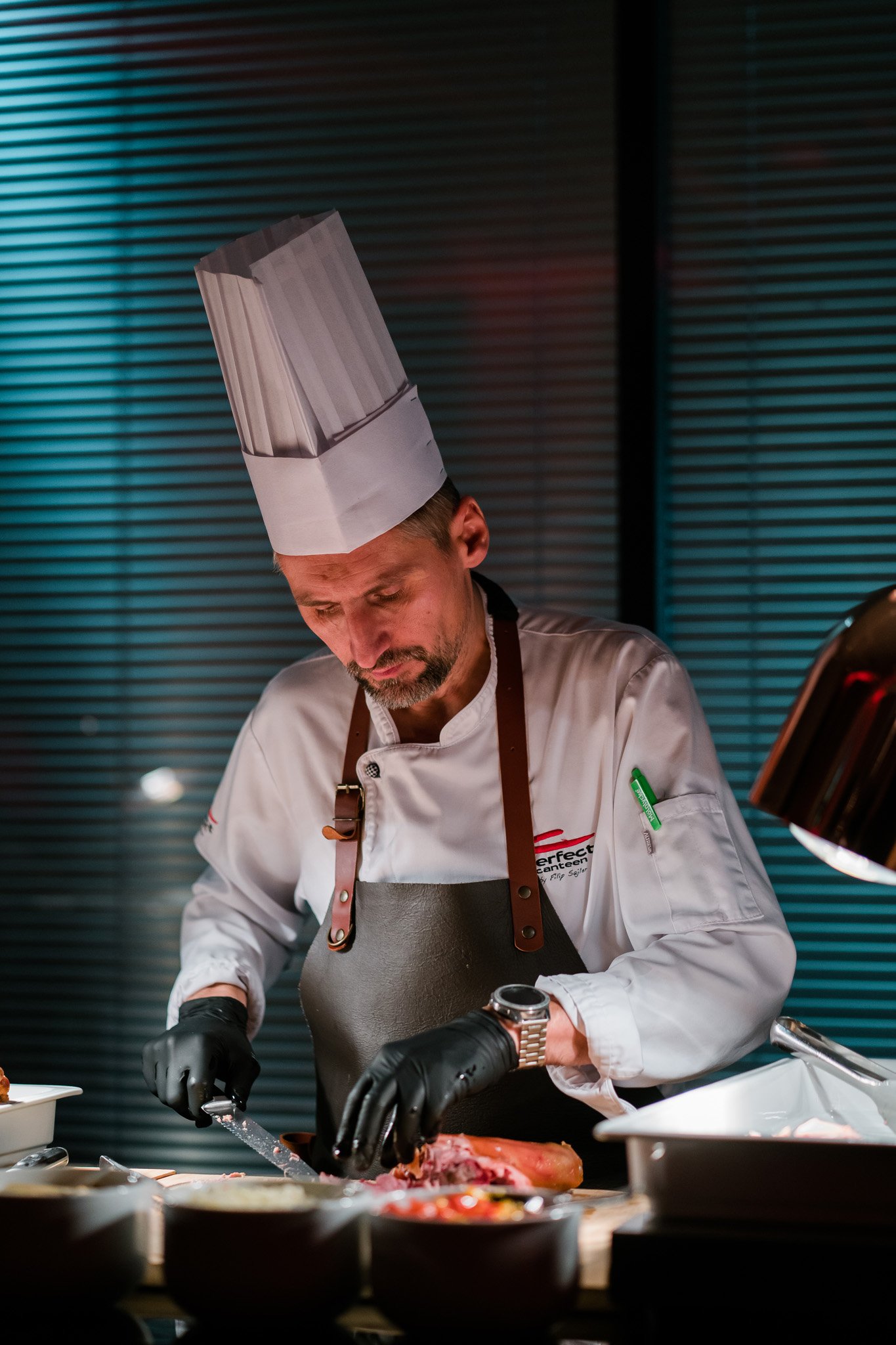 A male chef wearing a tall white chef hat, white chef jacket, and black gloves, is slicing meat on a cutting board in a professional kitchen. Catering and food photography in Zürich, Zug, Bern, Lucerne, Basel, St. Gallen and across Switzerland.