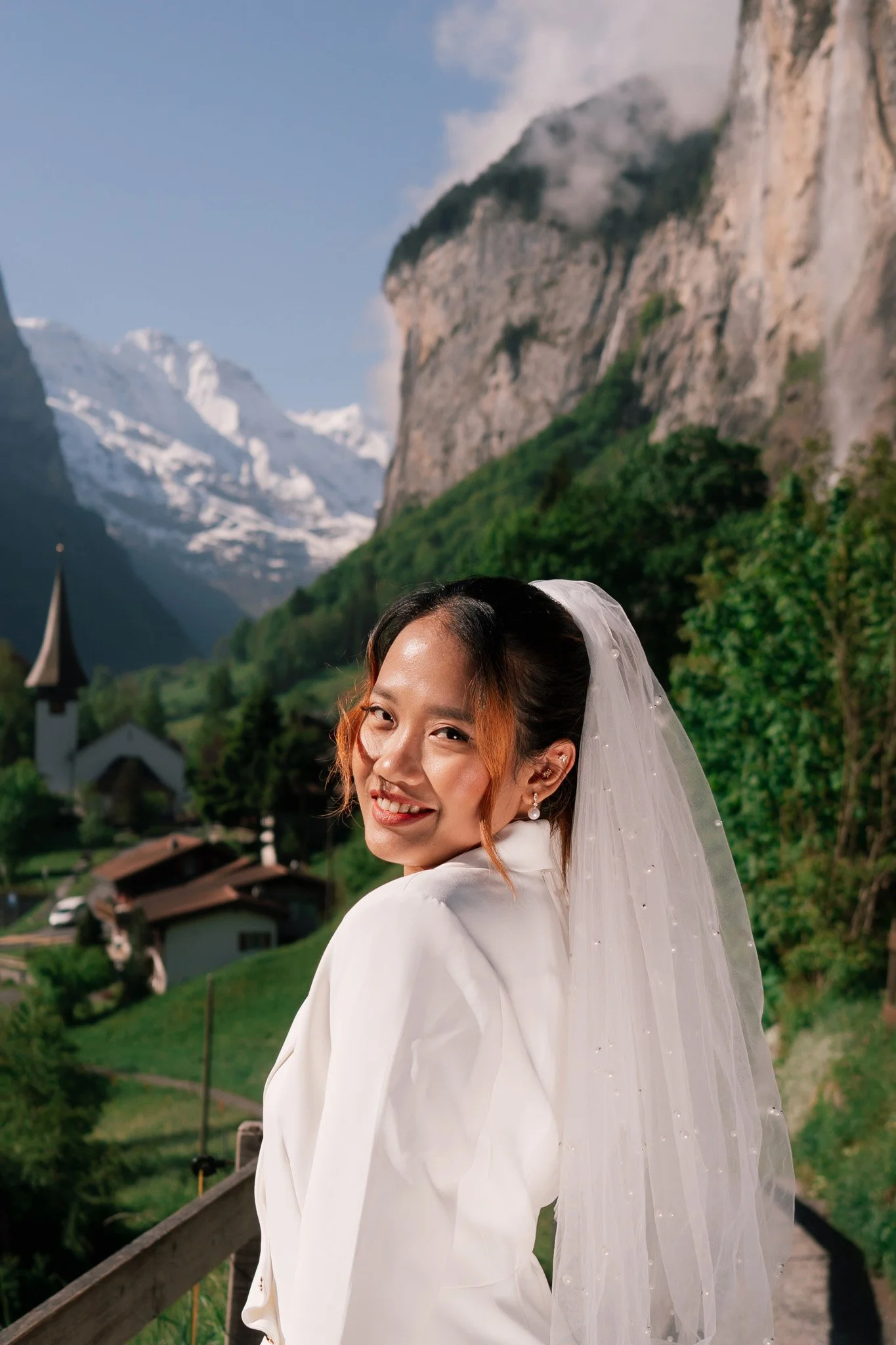 A smiling woman in wedding attire with a veil, standing outdoors with a mountainous and forested landscape in the background - Lauterbrunnen valley Switzerland.