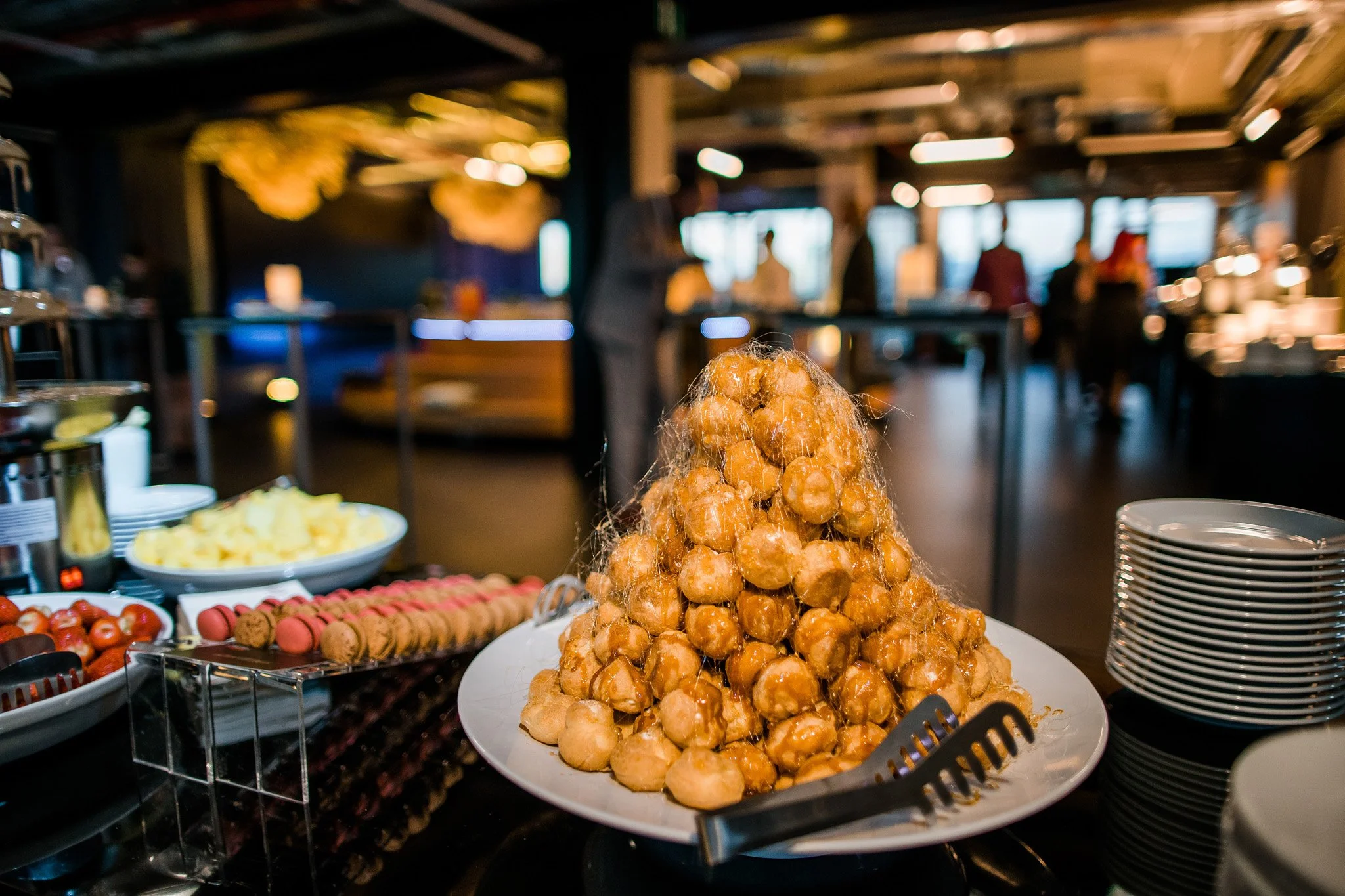 A pile of caramel-coated profiteroles on a white platter at a dessert buffet with plates and other desserts in the background. Catering, culinary and food photography in Zürich, Zug, Bern, Lucerne, Basel, St. Gallen and across Switzerland.