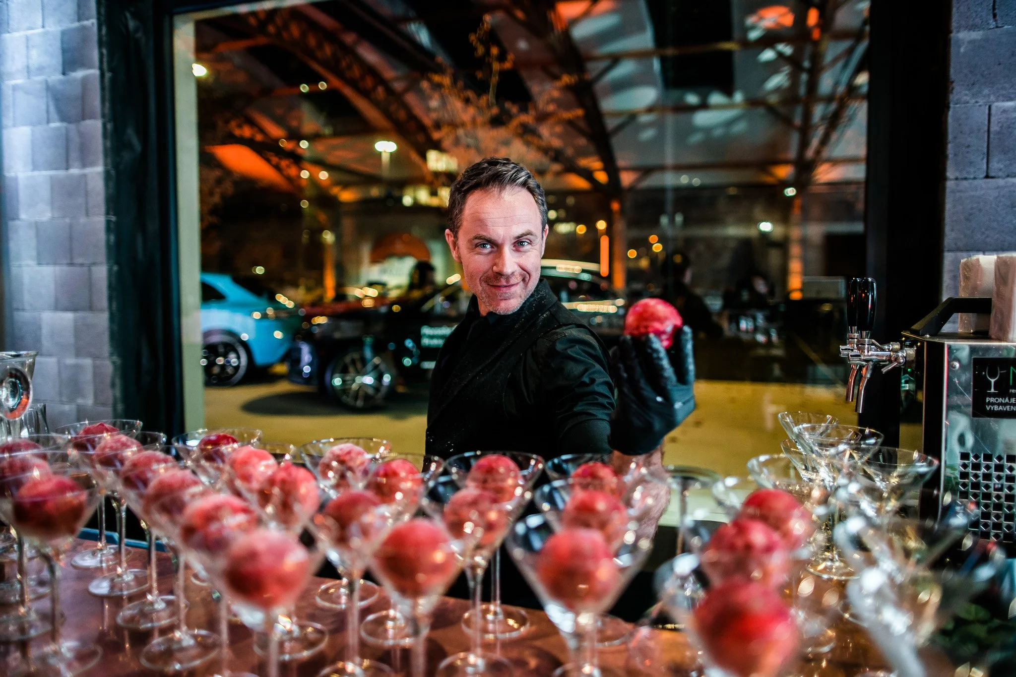 A man in black attire pouring drinks behind a bar surrounded by glass cups with pink-colored desserts. Catering and food photography in Zürich, Zug, Bern, Lucerne, Basel, St. Gallen and across Switzerland.