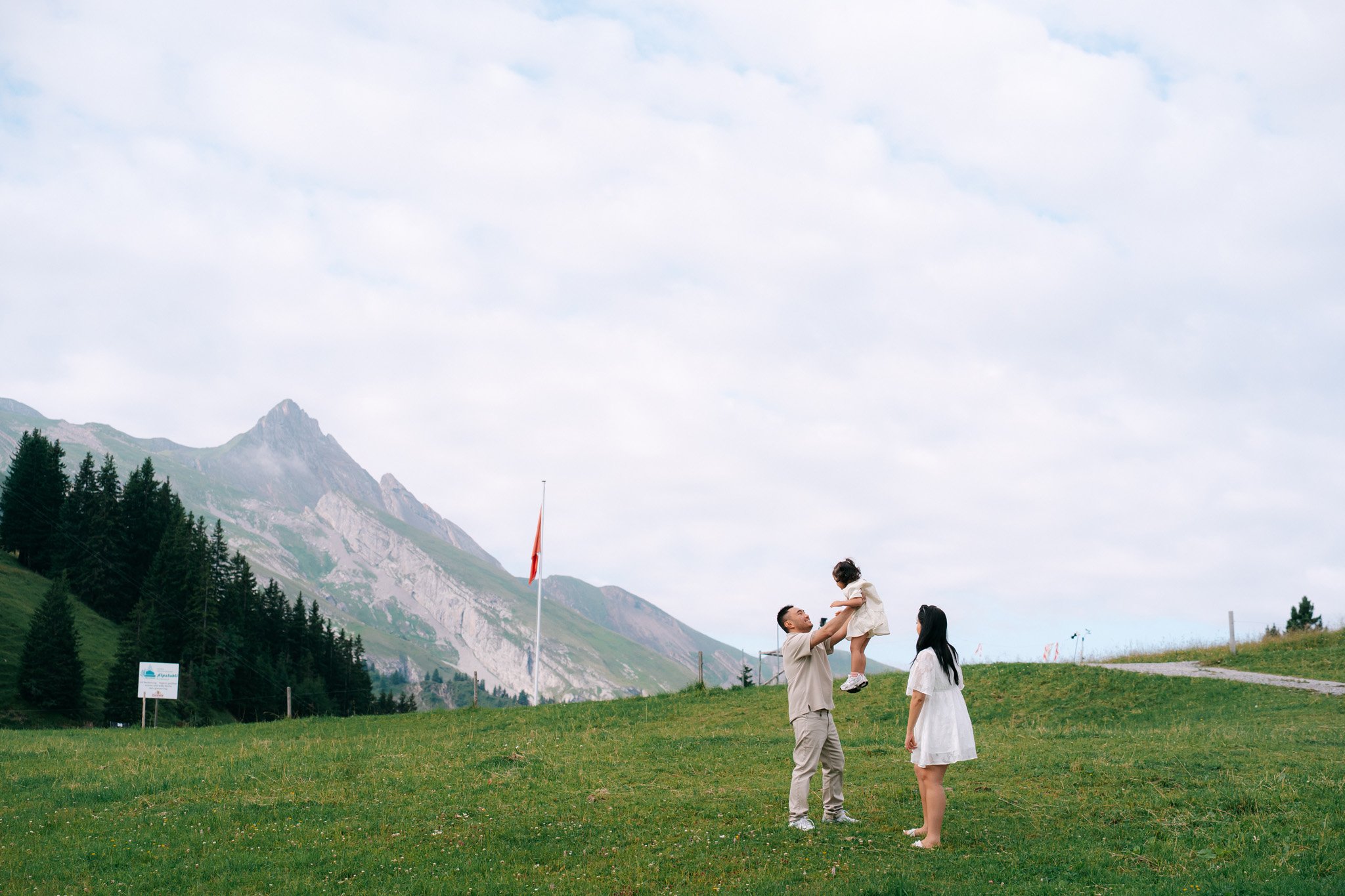 Family of four standing on a grassy hill with mountains in the background. The father is lifting a young girl, the mother and a teenage girl stand nearby, smiling. Family photoshoot in Switzerland.