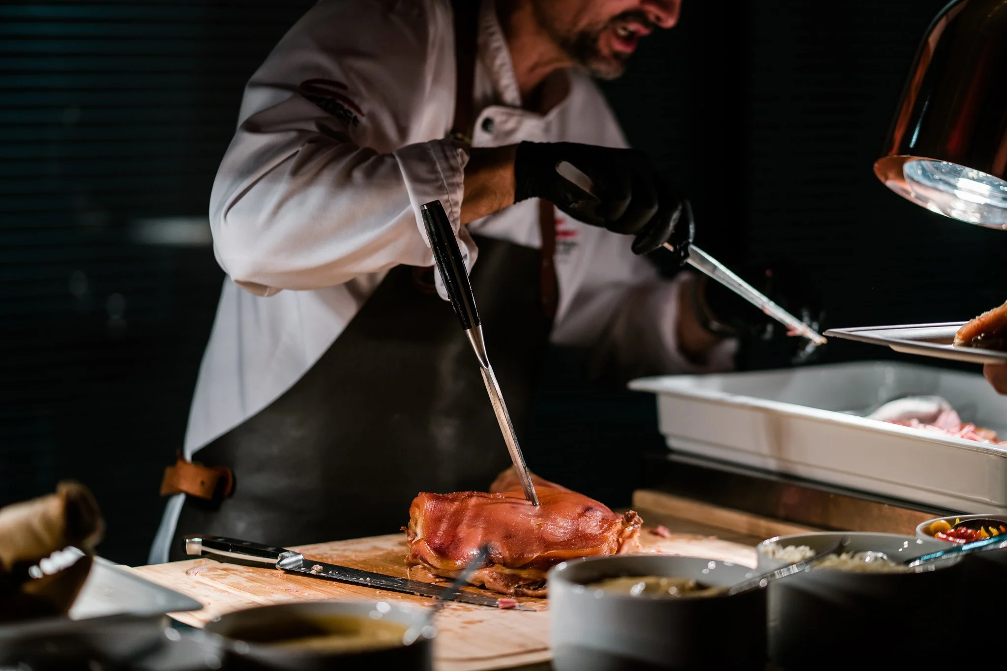 Chef in white coat and black gloves carving a large piece of ham on a wooden board in a professional kitchen. Catering and food photography in Zürich, Zug, Bern, Lucerne, Basel, St. Gallen and across Switzerland.