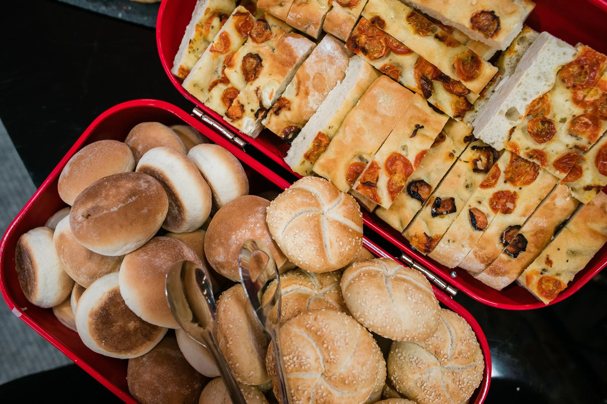 Two red trays filled with bread and baked goods. One tray contains small buns with some toasted tops, and round rolls with sesame seeds on top. Catering and food photography in Zürich, Zug, Bern, Lucerne, Basel, St. Gallen and across Switzerland.