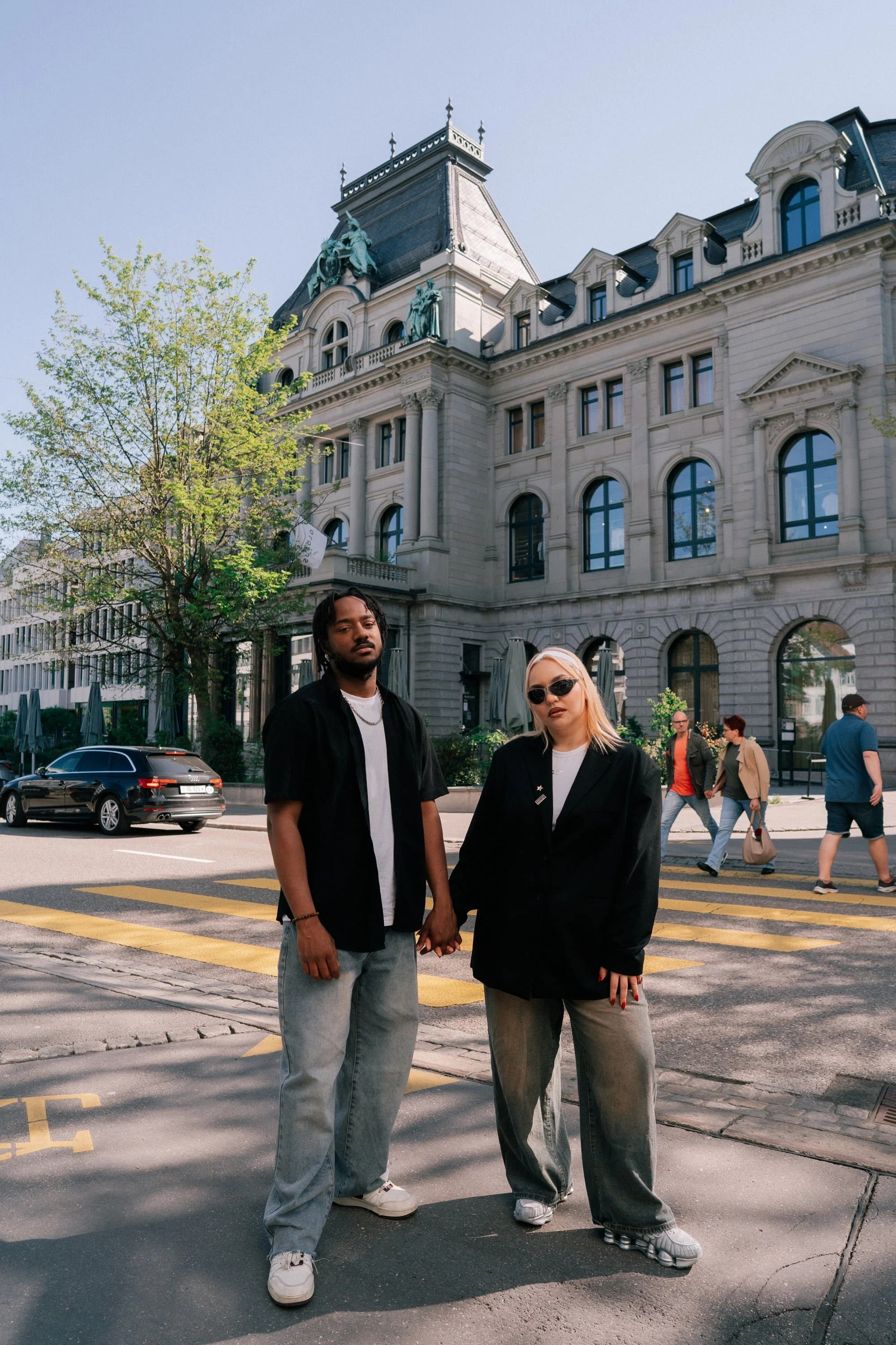 A man and woman holding hands standing on a city sidewalk in front of a large historic building with ornate architecture. The woman is wearing sunglasses and oversized clothing, and the man is dressed casually. Other pedestrians walk in the backgroun