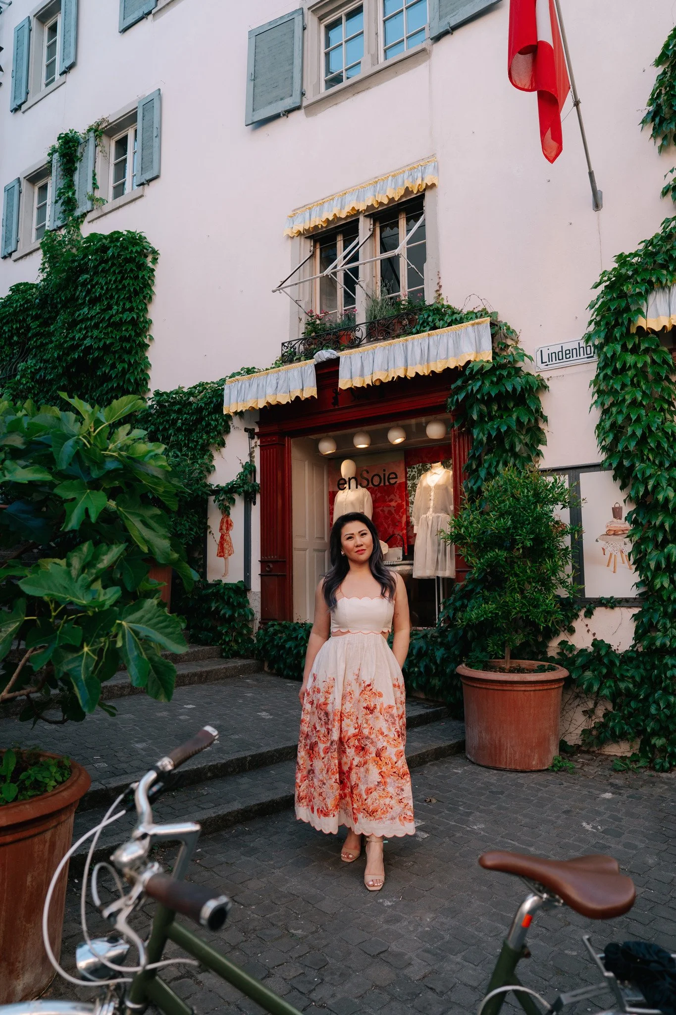 A woman in a floral dress standing outside a boutique called enSoie on Lindenhof street, with green ivy on the building wall, bicycles in the foreground. Solo traveler vacationing in Zurich.