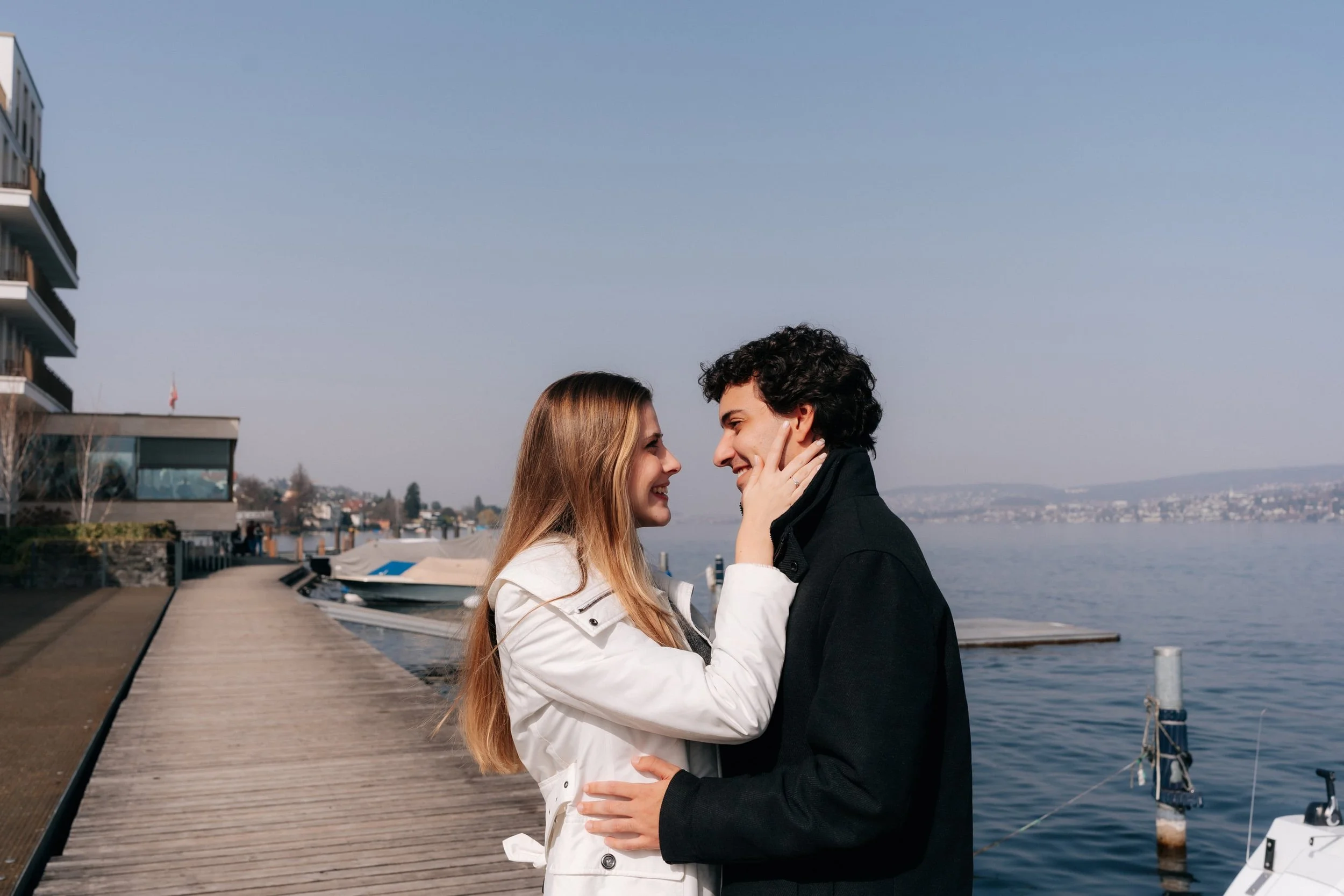 A young couple standing closely together on a wooden dock by a body of water, smiling and looking into each other's eyes, with boats and a shoreline in the background celebrating their surprise proposal in Switzerland. 