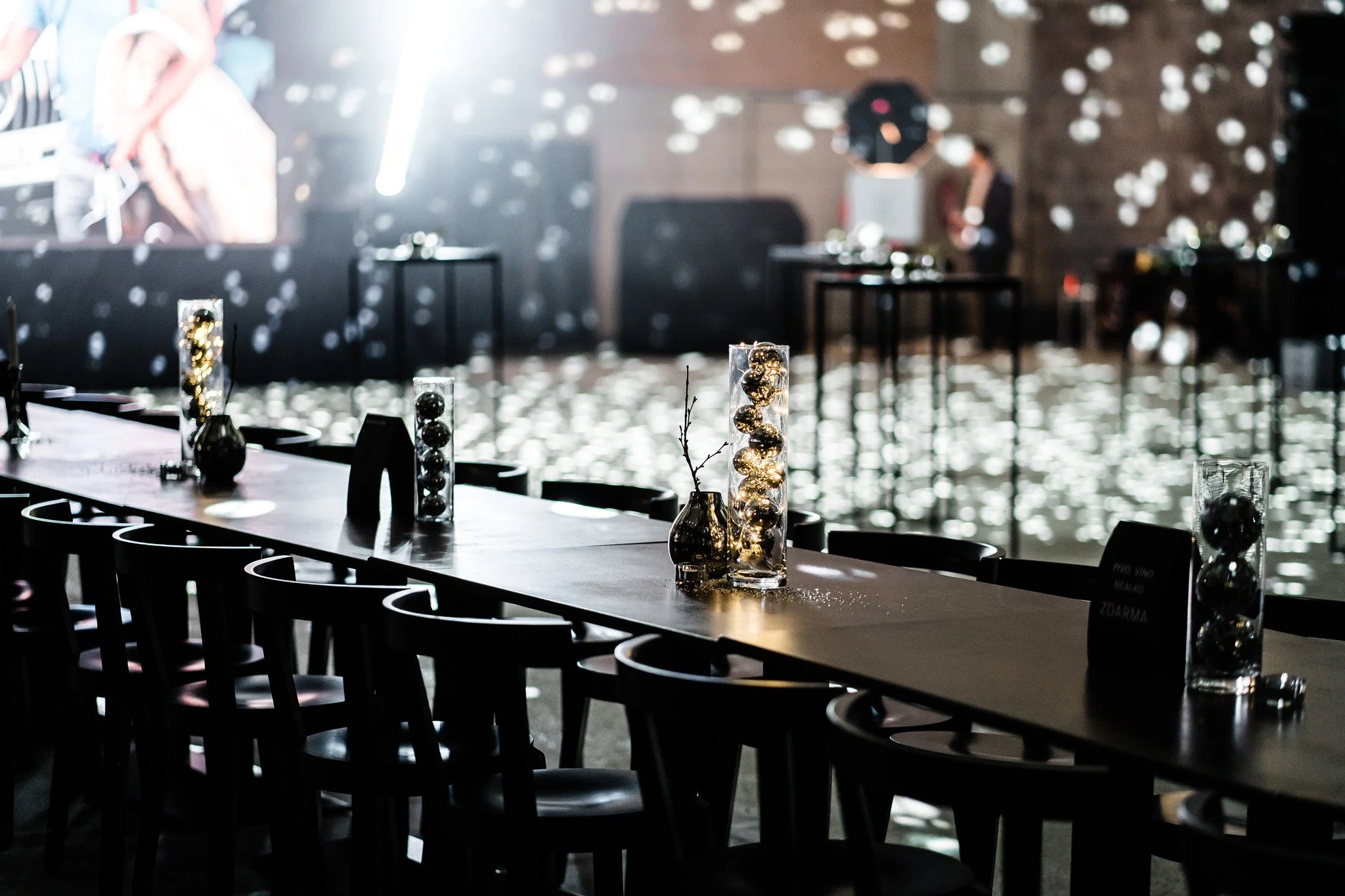 Event space with long table decorated with glass vases holding gold ornaments, illuminated by soft lighting, with tables and chairs, and a blurred background of a stage with a DJ setup and snowflake-like light reflections. Catering Switzerland.