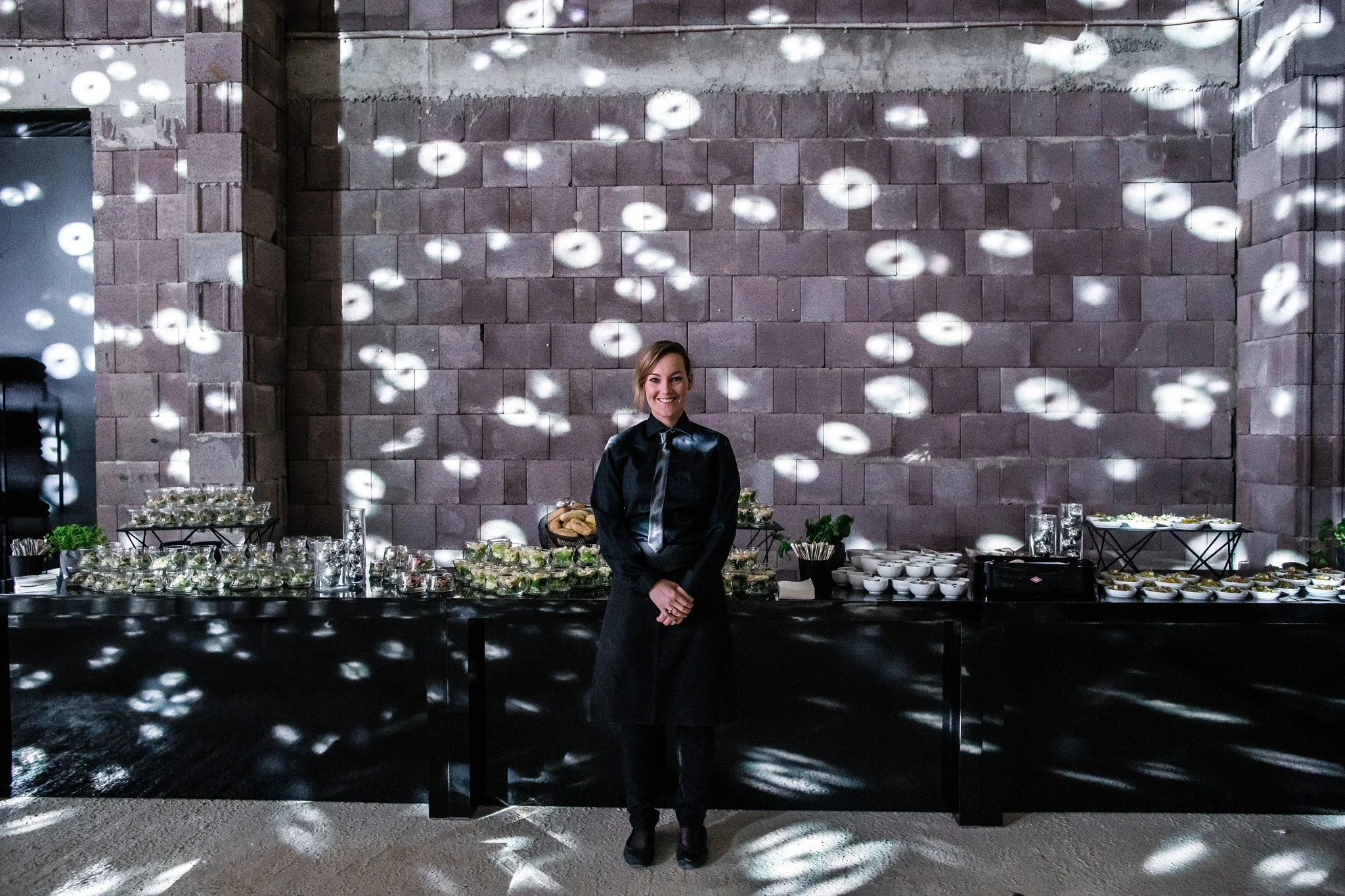 A smiling woman in black attire stands behind a dessert table in a dimly lit room with a brick wall background. The table is decorated. Catering and food photography in Zürich, Zug, Bern, Lucerne, Basel, St. Gallen and across Switzerland.