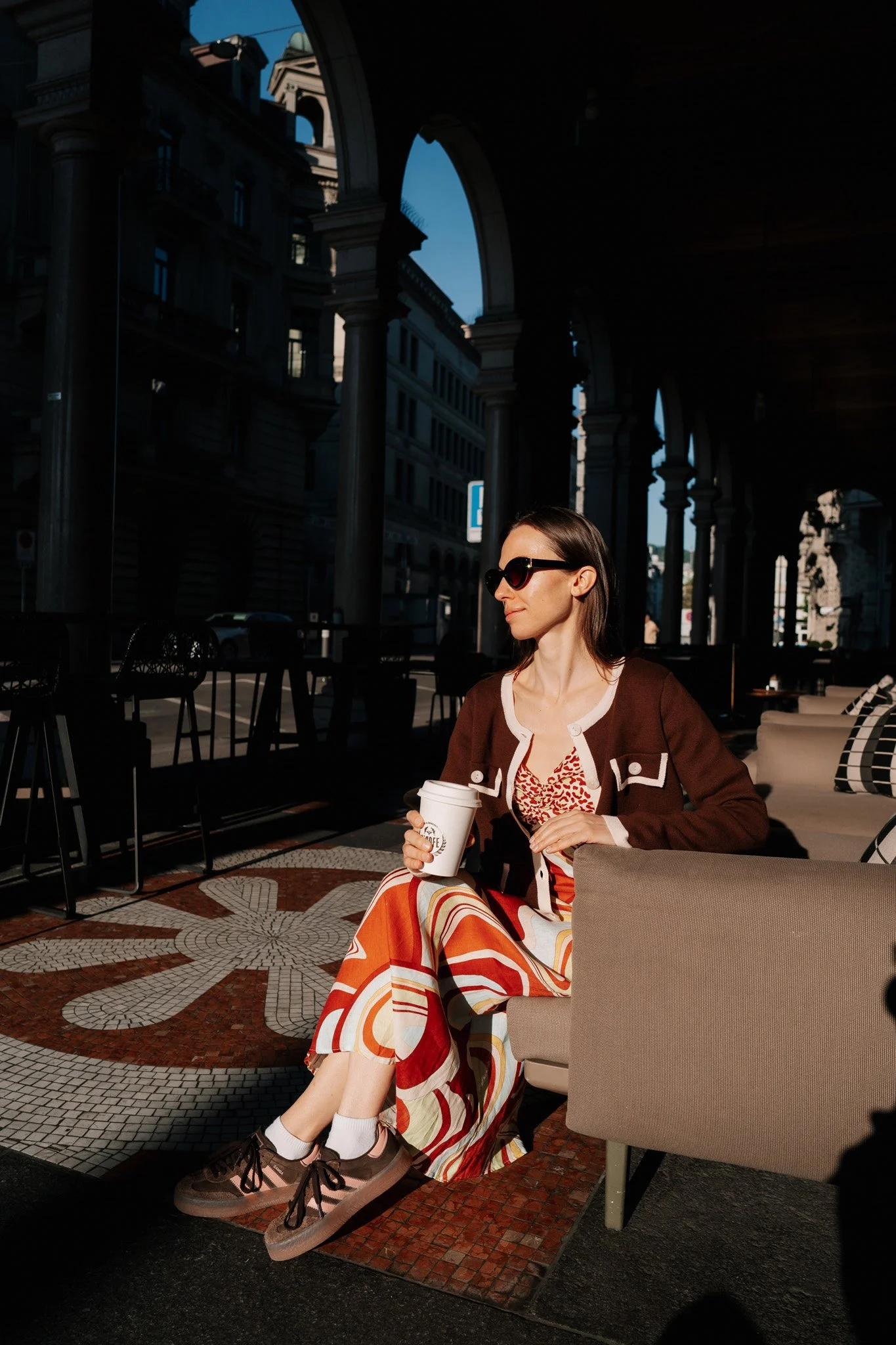 A woman wearing sunglasses in Zurich, a patterned dress, and a brown cardigan sitting in a cafe, holding a coffee cup, with tall arched buildings and a blue sky in the background.