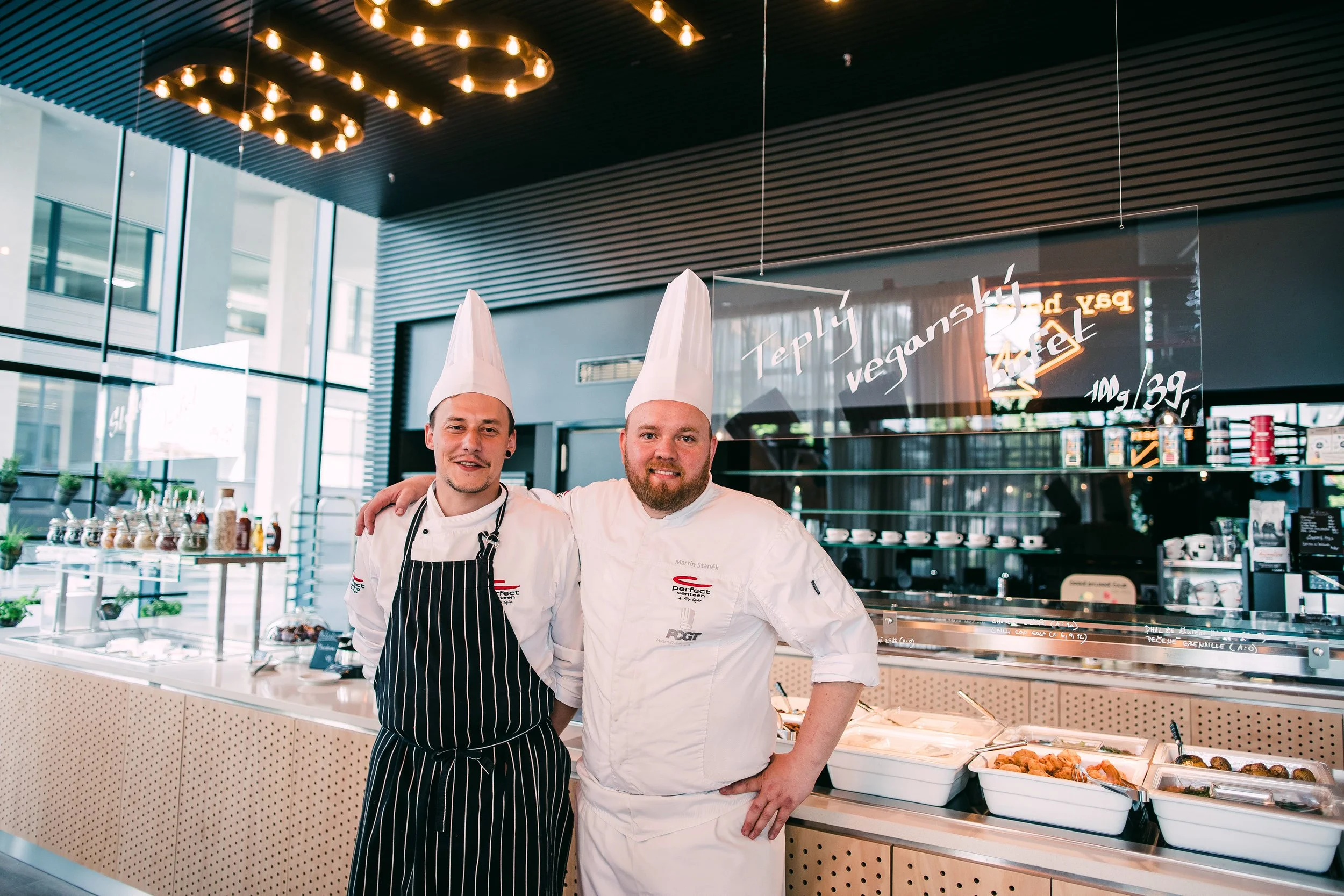 Two chefs in white uniforms and tall hats, standing side by side with arms around each other, smiling in a modern cafe or restaurant setting. Restaurant interior photography in Zürich, Zug, Bern, Lucerne, Basel, St. Gallen and across Switzerland.