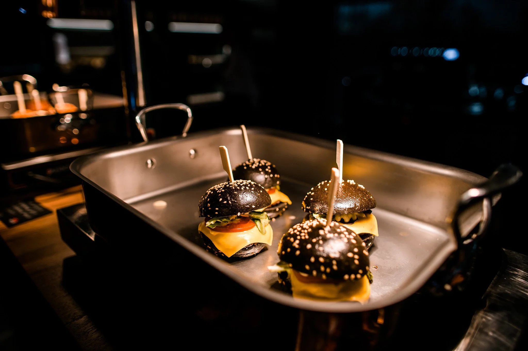 Mini black burger sliders with cheese and toppings on skewers in a metallic tray. Catering, culinary and food photography in Zürich, Zug, Bern, Lucerne, Basel, St. Gallen and across Switzerland.