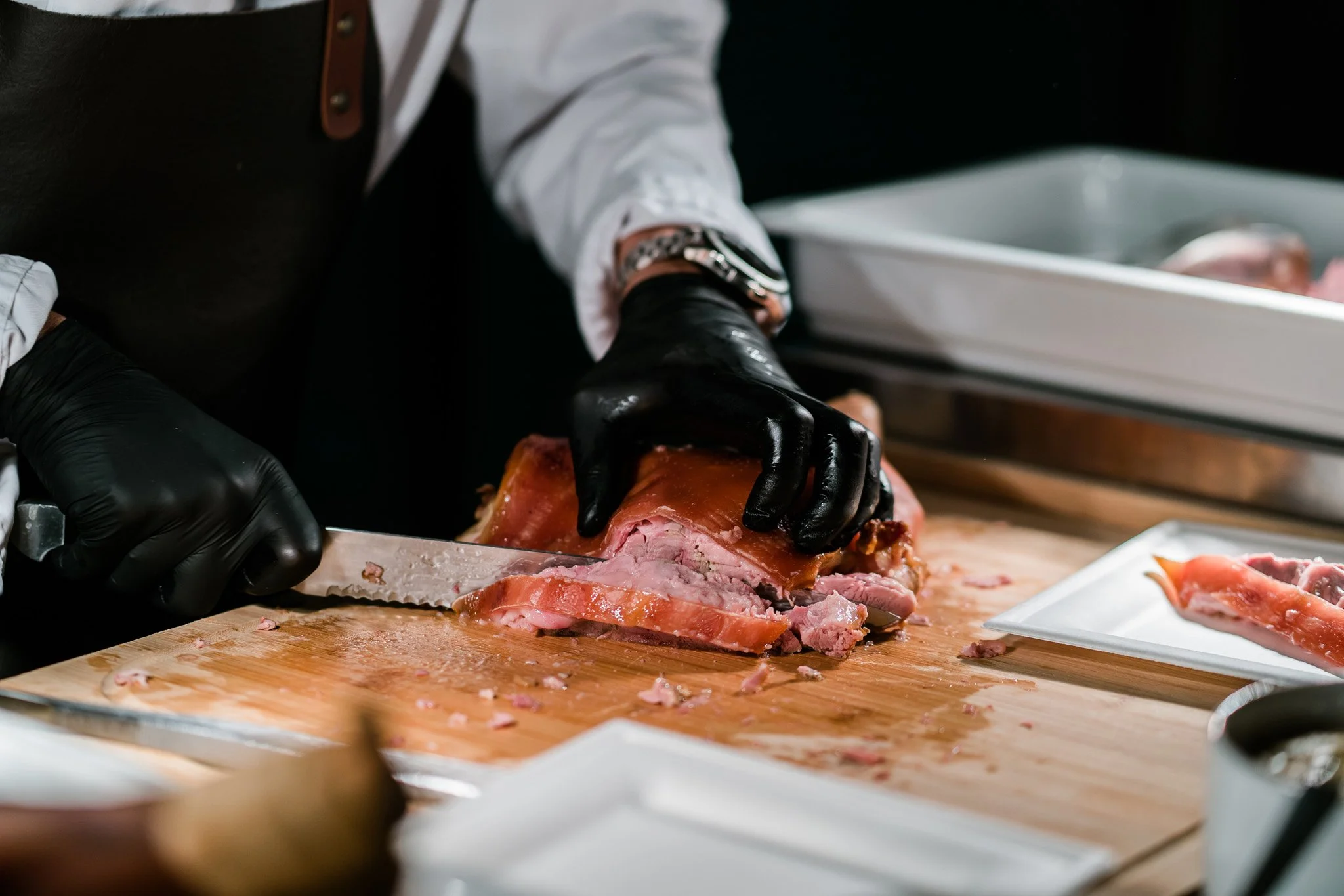 Person slicing cooked ham on a wooden cutting board, wearing black gloves and a white shirt. Catering, culinary and food photography in Zürich, Zug, Bern, Lucerne, Basel, St. Gallen and across Switzerland.