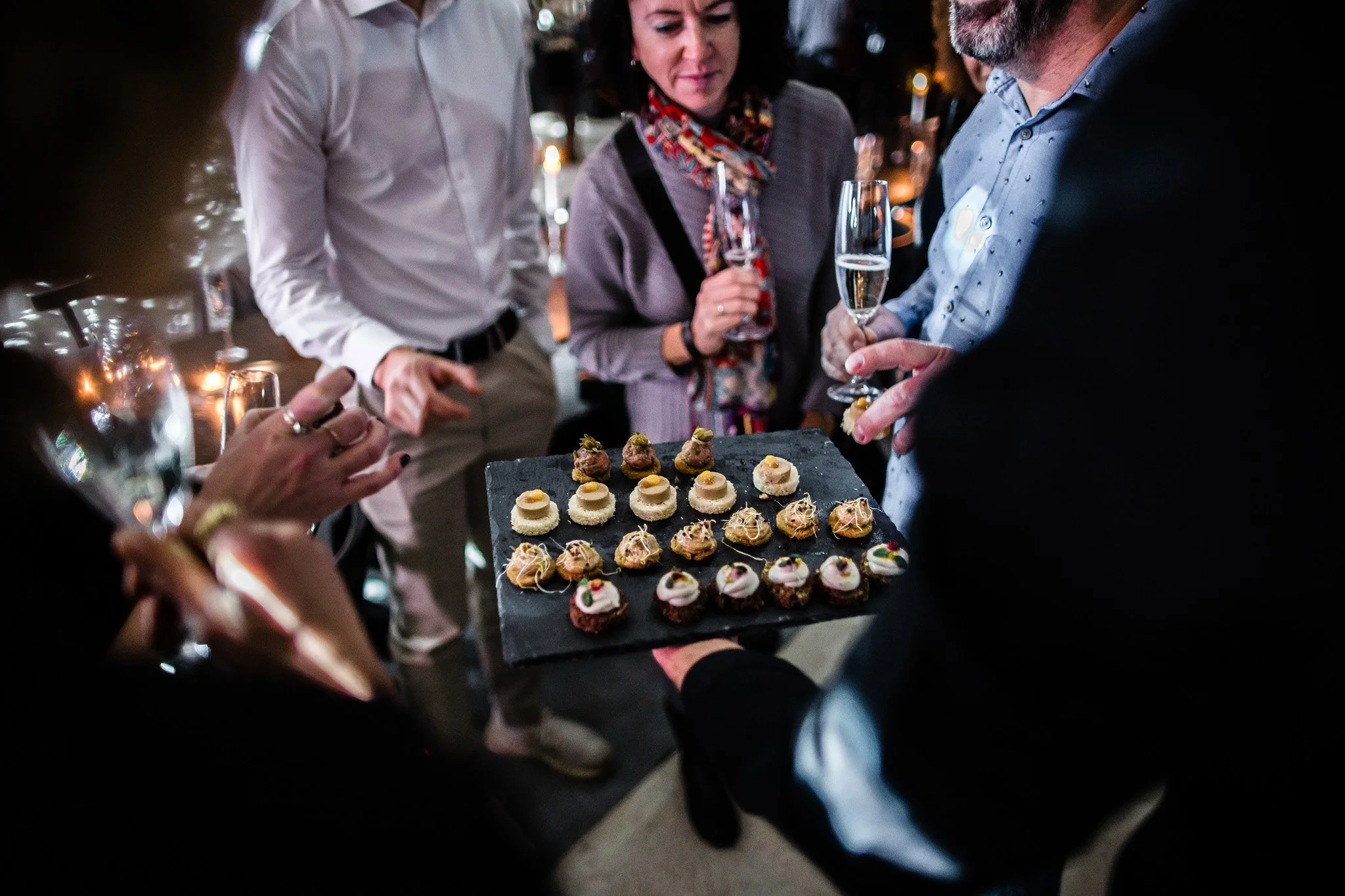 Group of people at a social gathering, standing around holding glasses of champagne, looking at a platter of assorted small appetizers or desserts. Catering, culinary and food photography in Zürich, Zug, Bern, Lucerne and across Switzerland.
