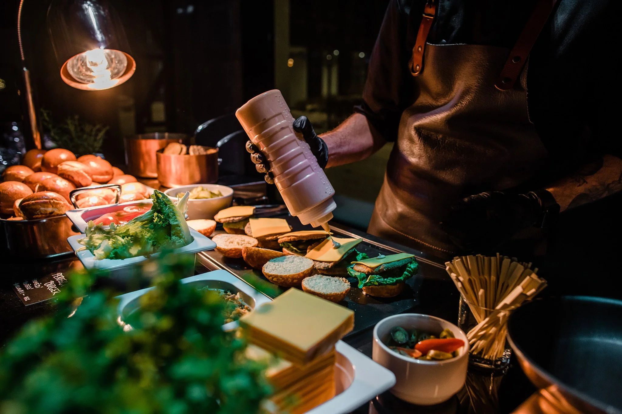 A person preparing sandwiches at a buffet table with various breads, lettuce, cheese, and condiments, under warm lighting. Catering, culinary and food photography in Zürich, Zug, Bern, Lucerne, Basel, St. Gallen and across Switzerland.