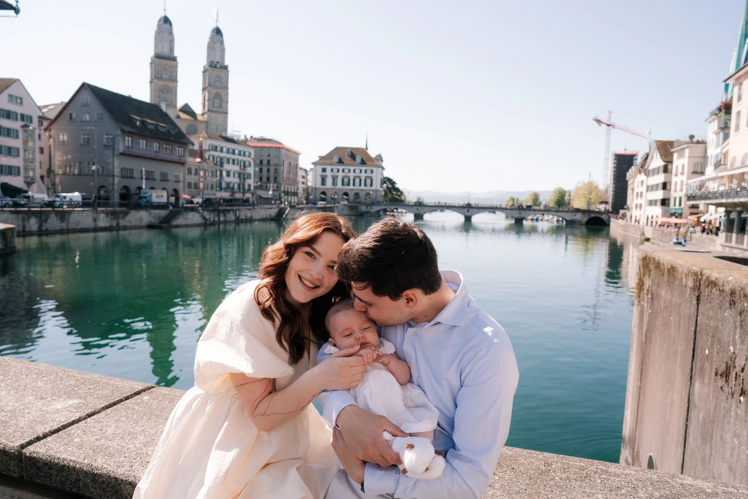 Family of three by river with bridge, historic buildings of Zurich Switzerland, church towers, and blue sky in the background