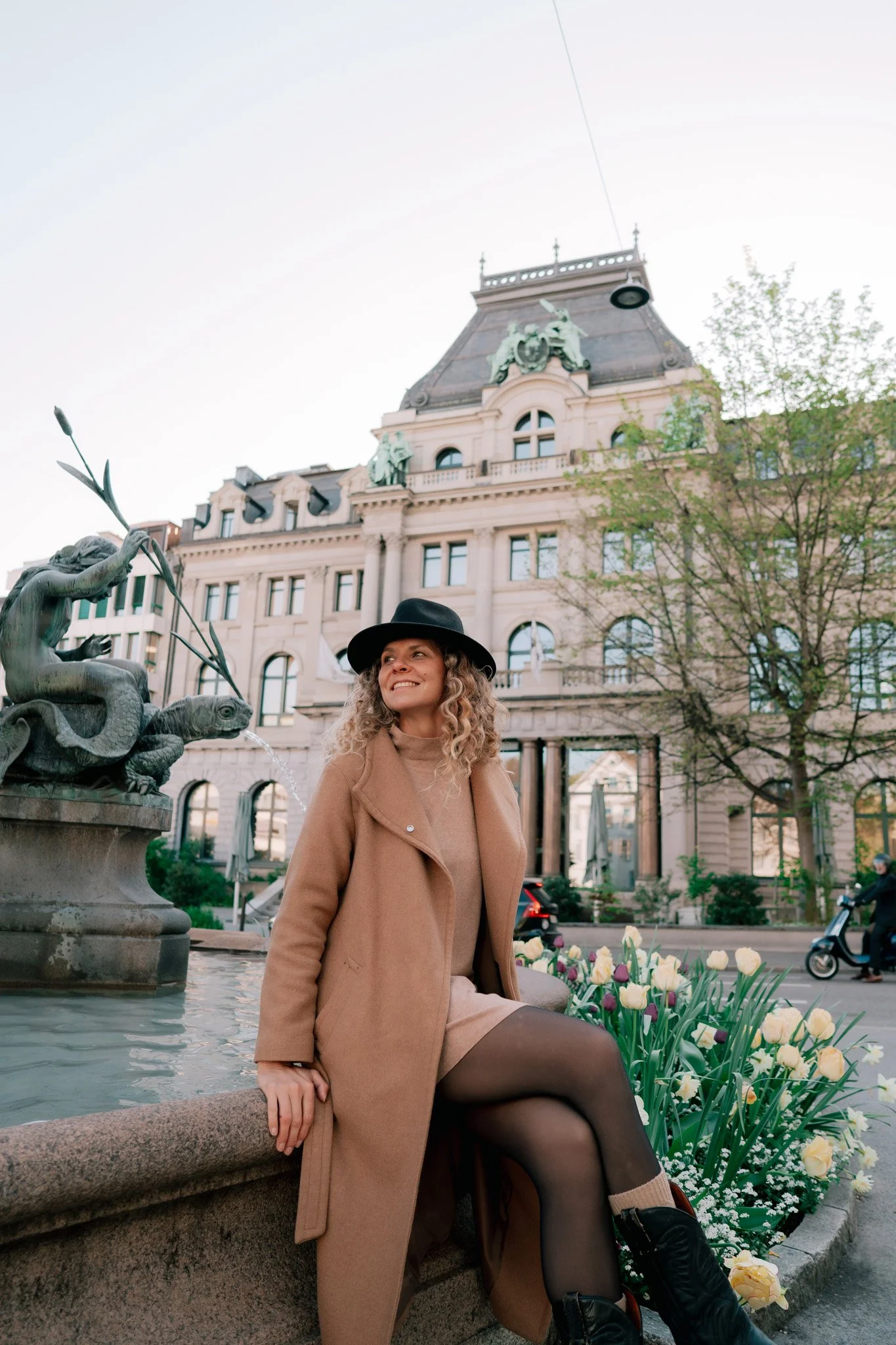 A woman sitting on the edge of a fountain in St. Gallen, Switzerland, with a building and trees in the background doing vacation photoshoot.