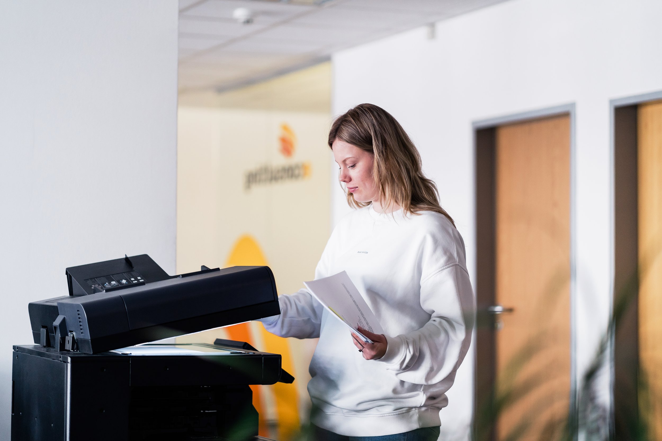 A woman with shoulder-length light brown hair in a white sweatshirt works at a black photocopy machine, holding papers in her left hand. The background shows a blurred office space with wooden doors and a white wall with a logo.
