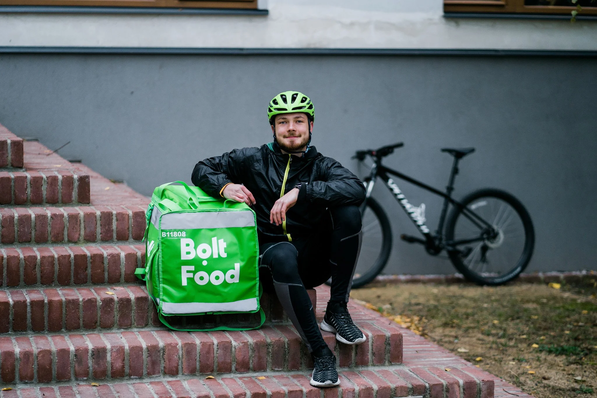 A man wearing a yellow helmet and black cycling attire sitting on brick steps next to a green Bolt Food delivery bag with a bicycle in the background. Advertising campaign photographer for corporate and startup business Switzerland.