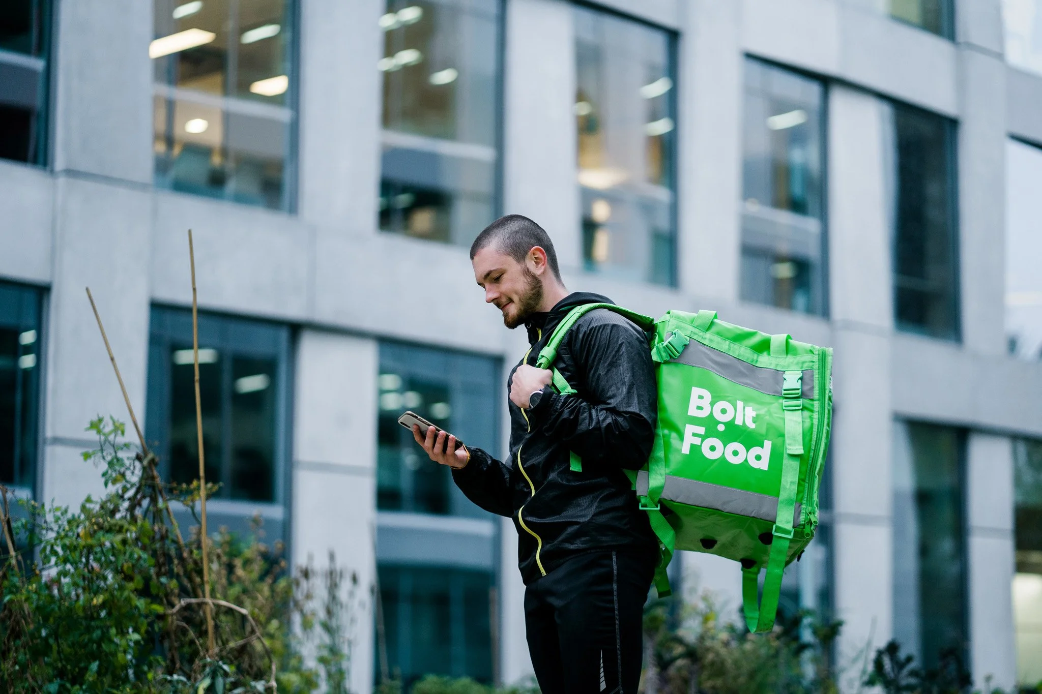 A man with a beard and short hair, wearing a black jacket, using a smartphone outdoors in front of a modern building. He has a large green Bolt Food delivery backpack. Advertising campaign photographer for corporate and startup business Switzerland.
