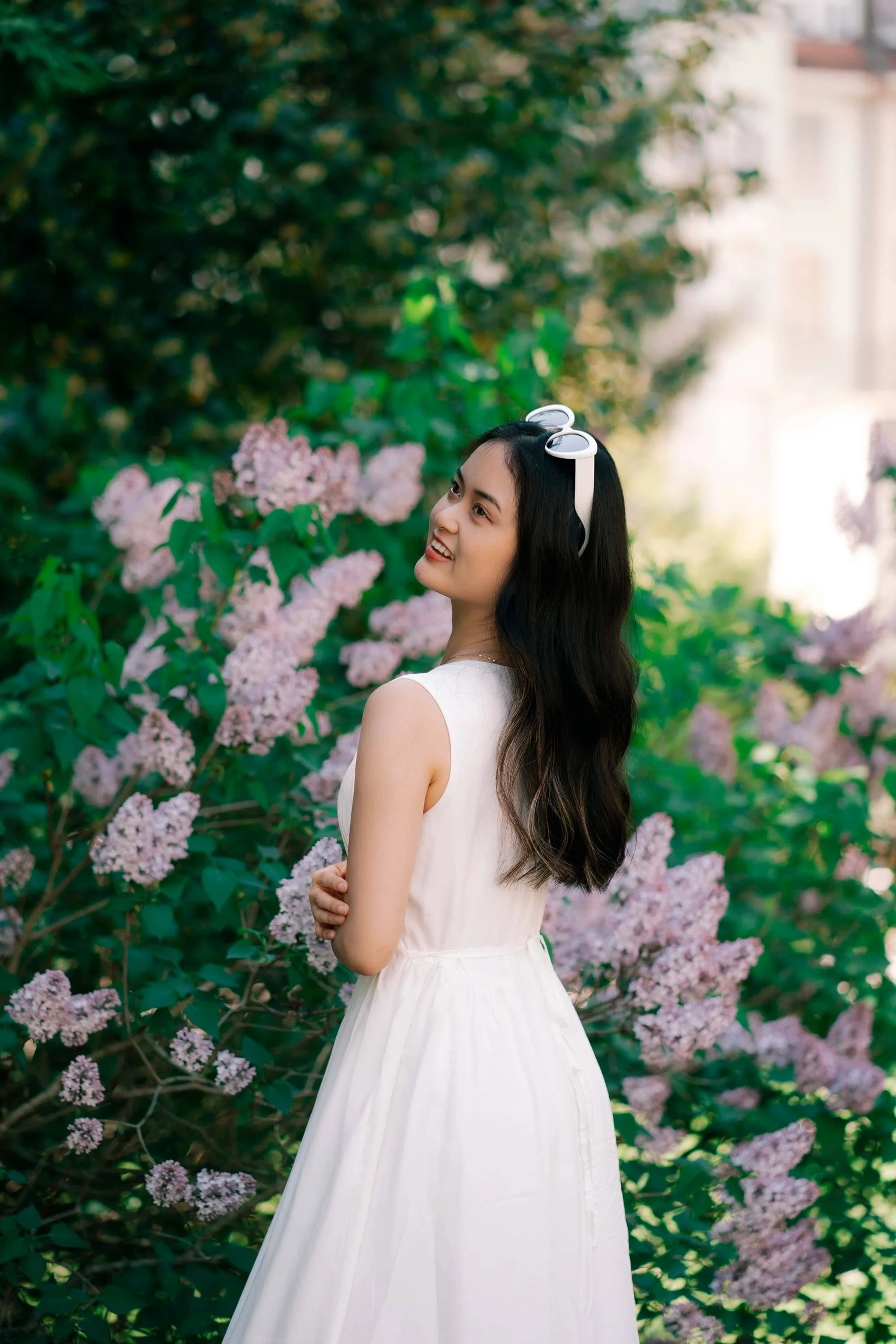 A woman with long black hair wearing a white dress and white sunglasses on her head, smiling and looking to the side, standing outdoors in front of pink flowering bushes and green foliage. Gardens in Zurich.