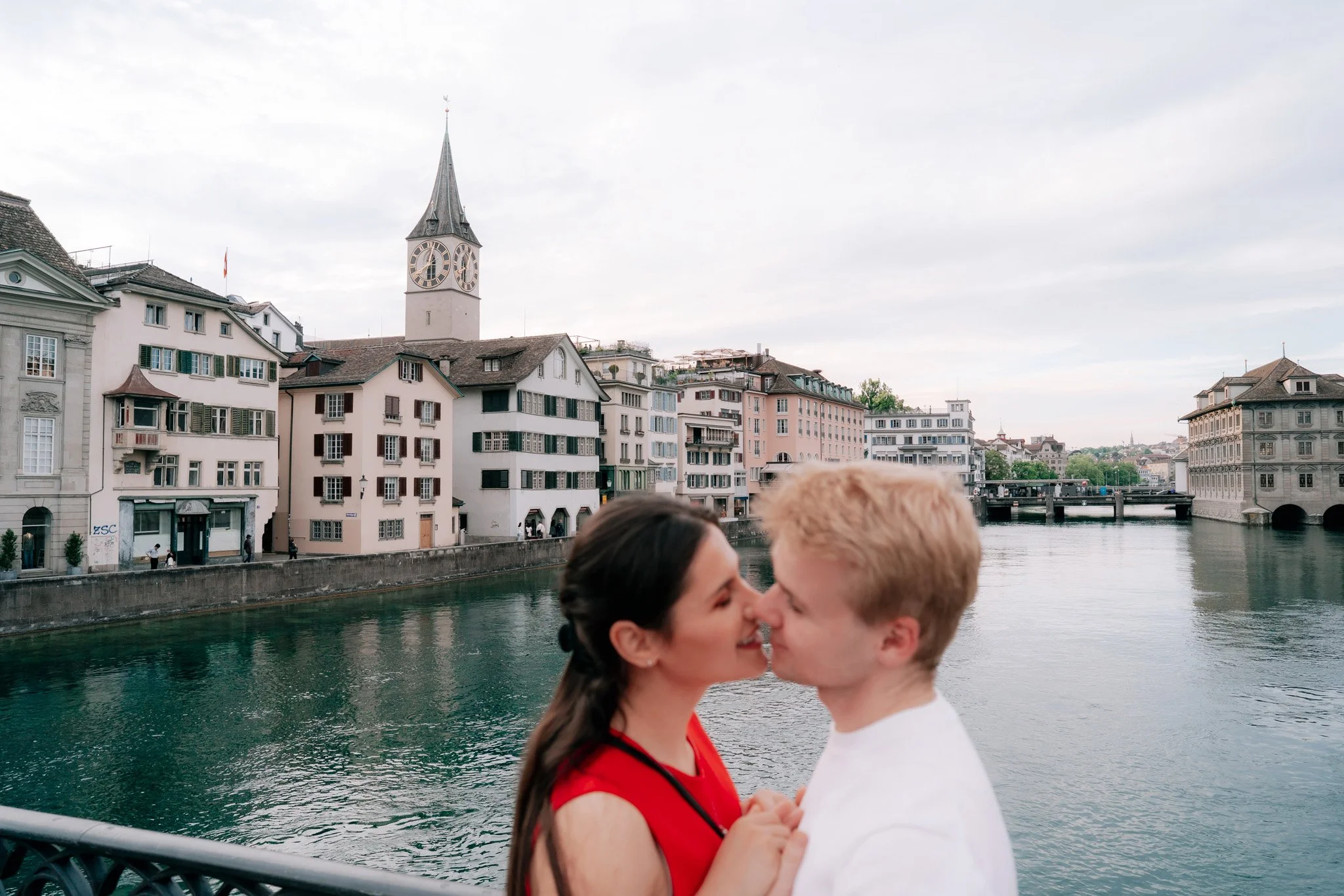 A couple kissing on a bridge over a river with historic buildings and a church steeple in the background, overlooking Zurich river.