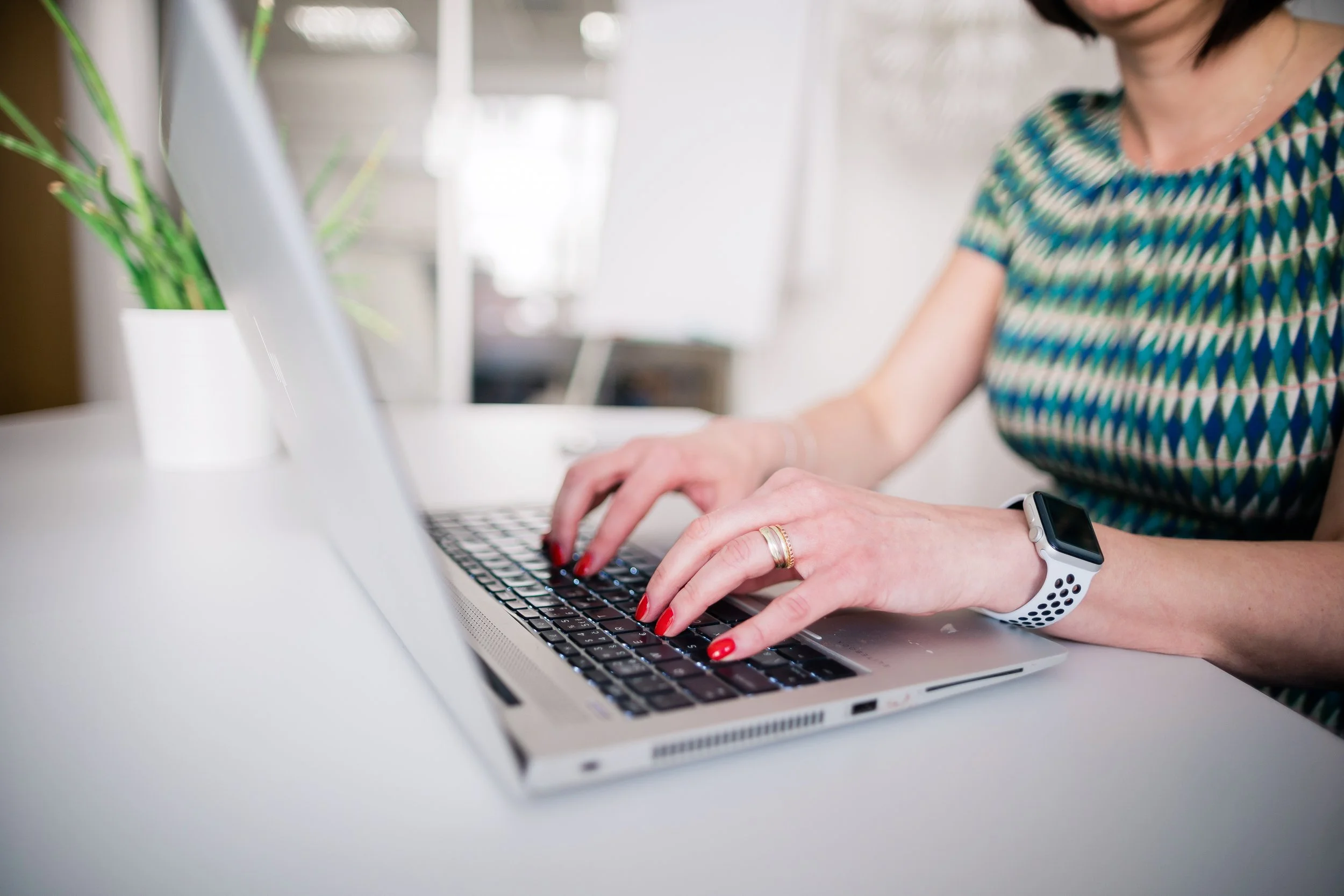 A woman typing on a silver laptop with a black keyboard, wearing a patterned blue and green dress and a white smartwatch, with a potted plant nearby. Corporate photography in Zürich, Zug, Bern, Lucerne, Basel, St. Gallen - across Switzerland.