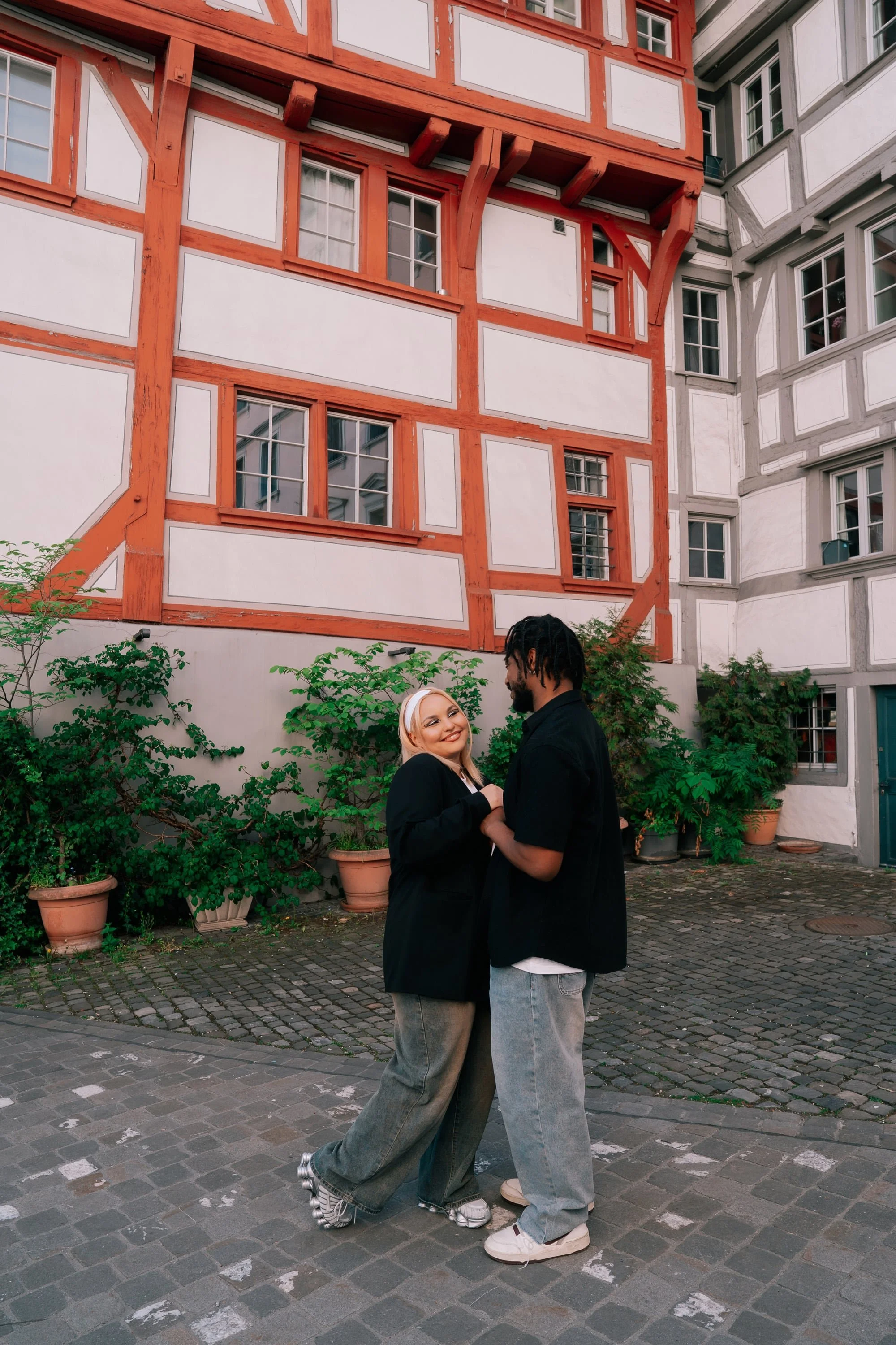 A couple holding hands and smiling at each other in a courtyard with cobblestone pavement, potted plants, and a building in St Gallen Switzerland with red and white timber framing.