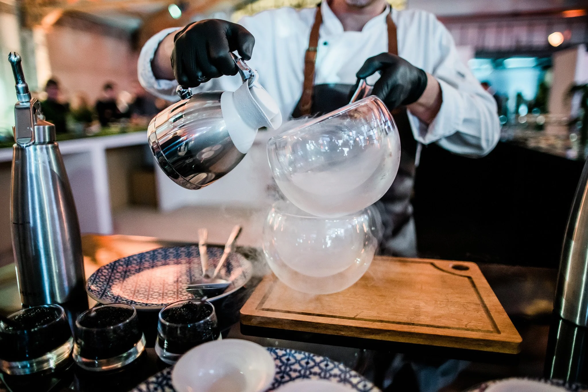A bartender in a white shirt and black gloves is pouring liquid from a metal kettle into a spherical glass. The setup includes a wooden board and various glasses and plates on the table. Catering and food photography in Switzerland.