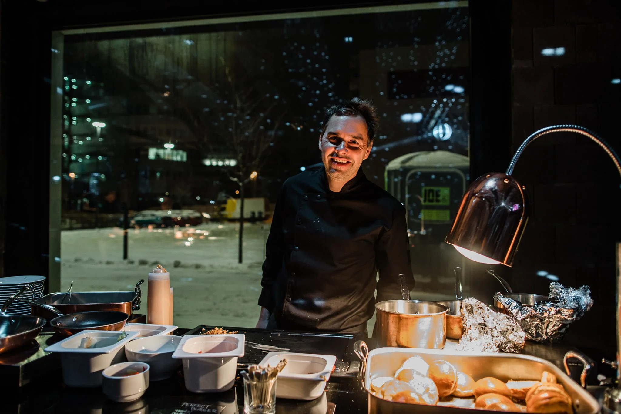 A smiling chef standing behind a counter with cooking utensils and ingredients, in a restaurant kitchen during nighttime, with a window showing a night cityscape. Catering and food photography in Zürich, Zug, Bern, Lucerne, Basel, St. Gallen.