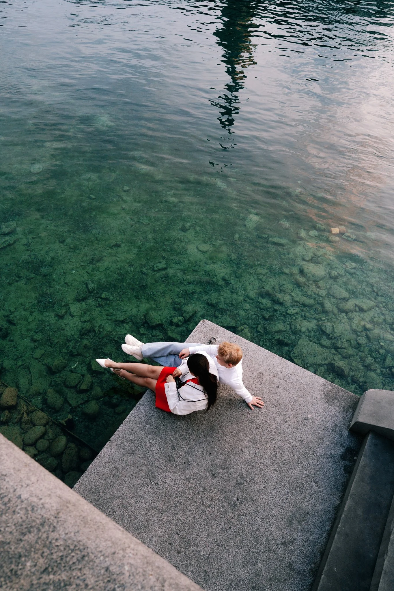 Two people sitting on a concrete ledge by the water, having romantic date in Zurich Switzerland, with one person resting their arm on the other. The water is clear and calm, showing rocks beneath the surface.