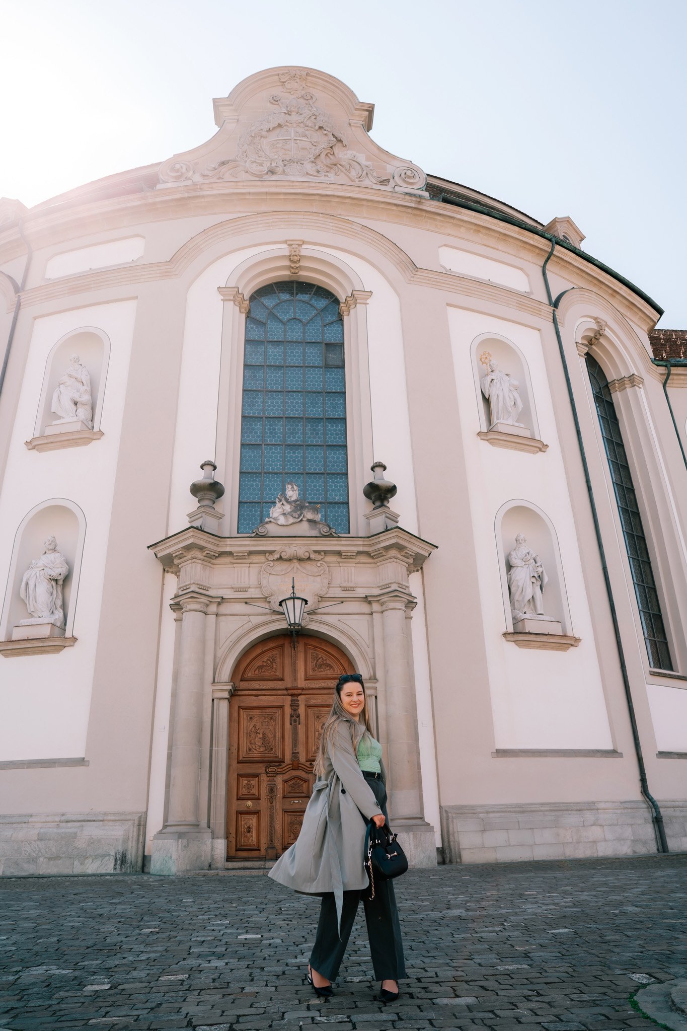 Solo traveler woman standing in front of a large, ornate church in St. Gallen, Switzerland, with statues in niches and a wooden door, smiling at the camera.