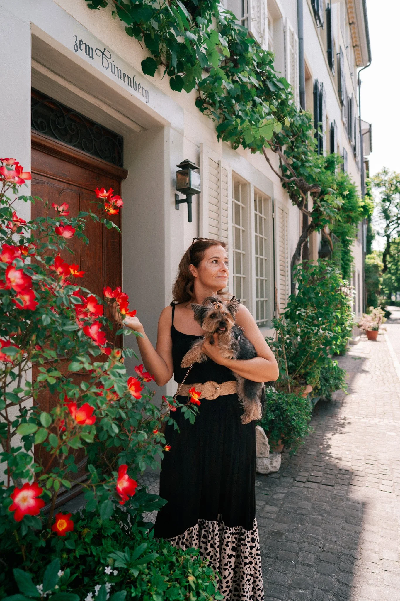 Vacation photoshoot of woman in black dress holding a small dog standing outdoors on a cobblestone street near a house with greenery and flowers in Basel, Switzerland.