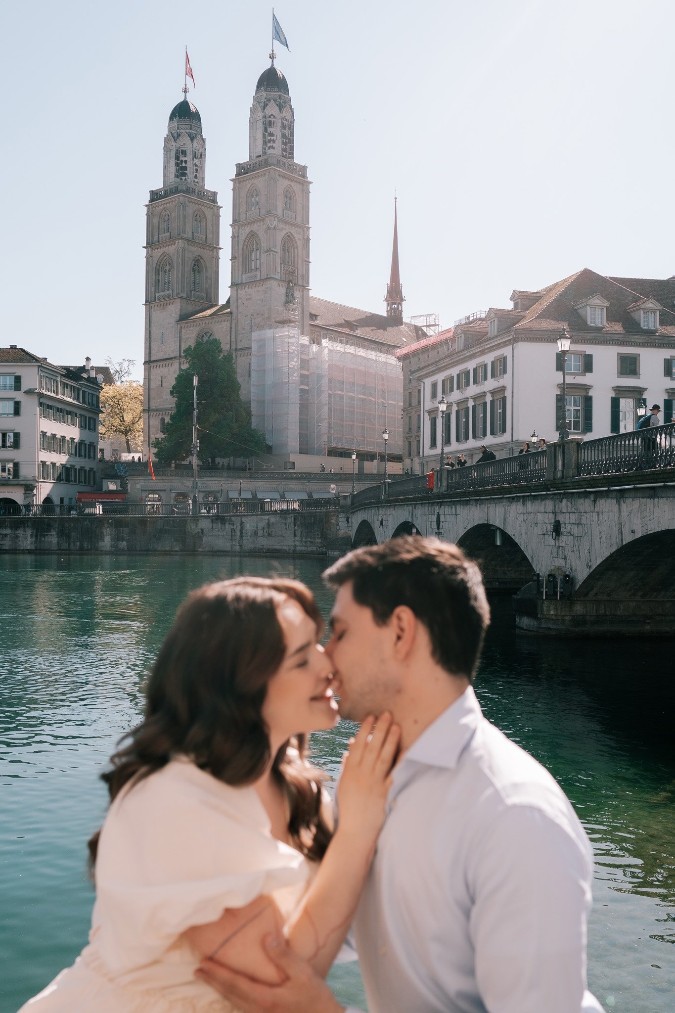 A romantic couple kissing in front of a river with a church and bridge in the background on a vacation in Switzerlands historic city of Zurich.