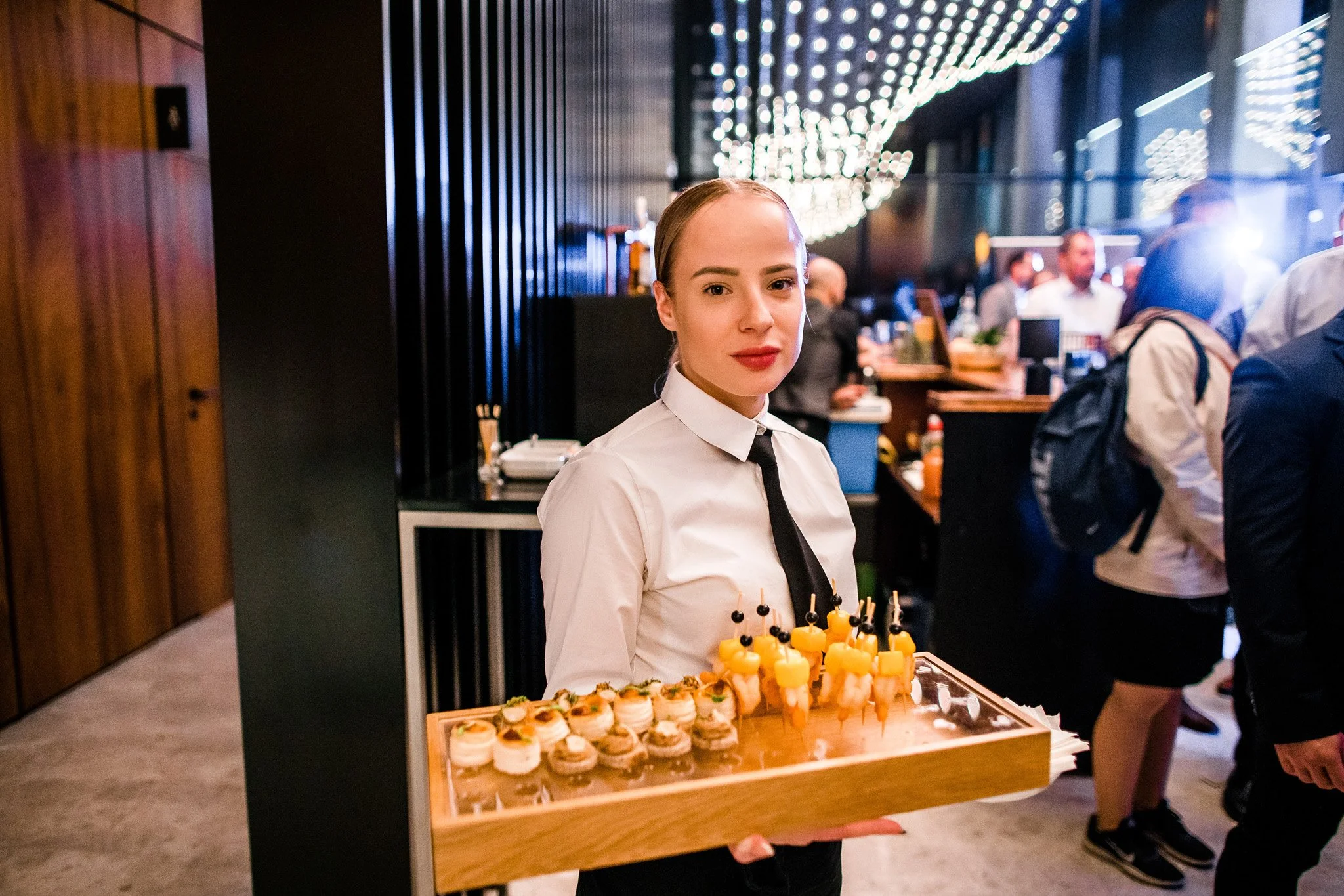 A young woman in a white shirt and black tie holding a tray of small appetizers at a crowded event or restaurant. Catering, culinary and food photography in Zürich, Zug, Bern, Lucerne, Basel, St. Gallen and across Switzerland.