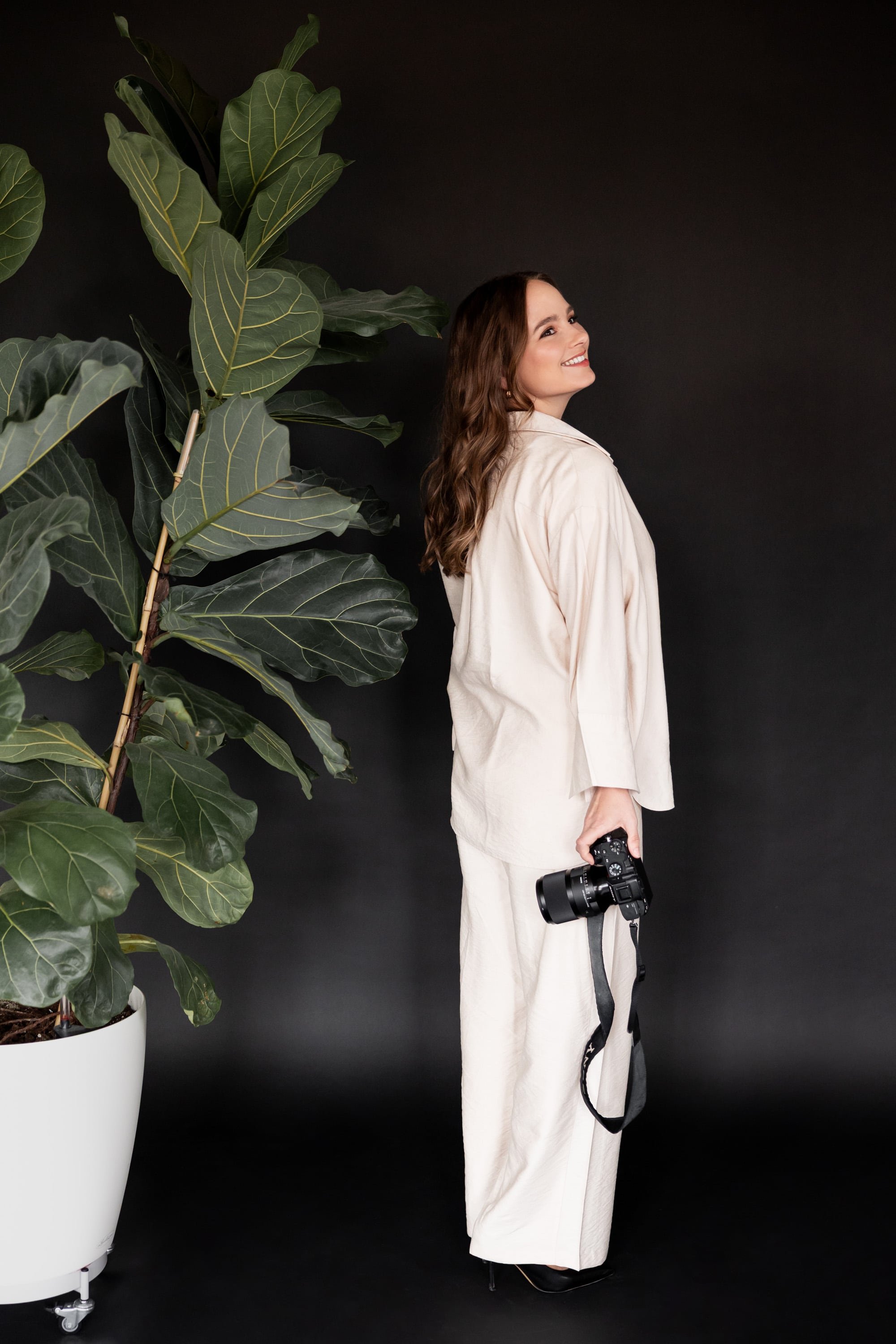 Switzerland vacation photographer in beige outfit holding a camera, standing next to a large potted plant against a dark background.