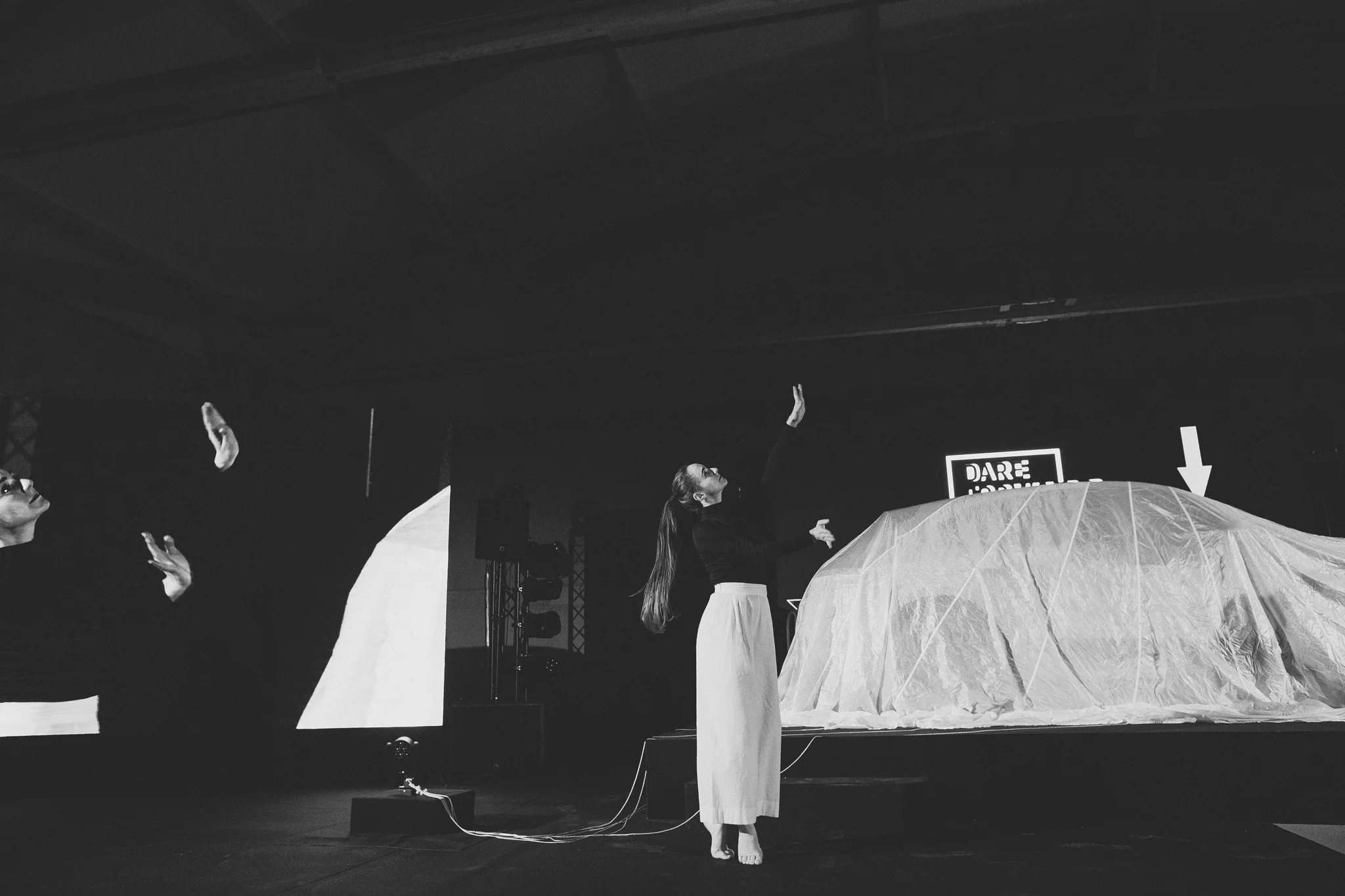 A woman with long hair, dressed in a black top and light-colored skirt, performs on stage with her arm raised towards the ceiling, while two other performers with glasses are to her left. Car event photography Switzerland.