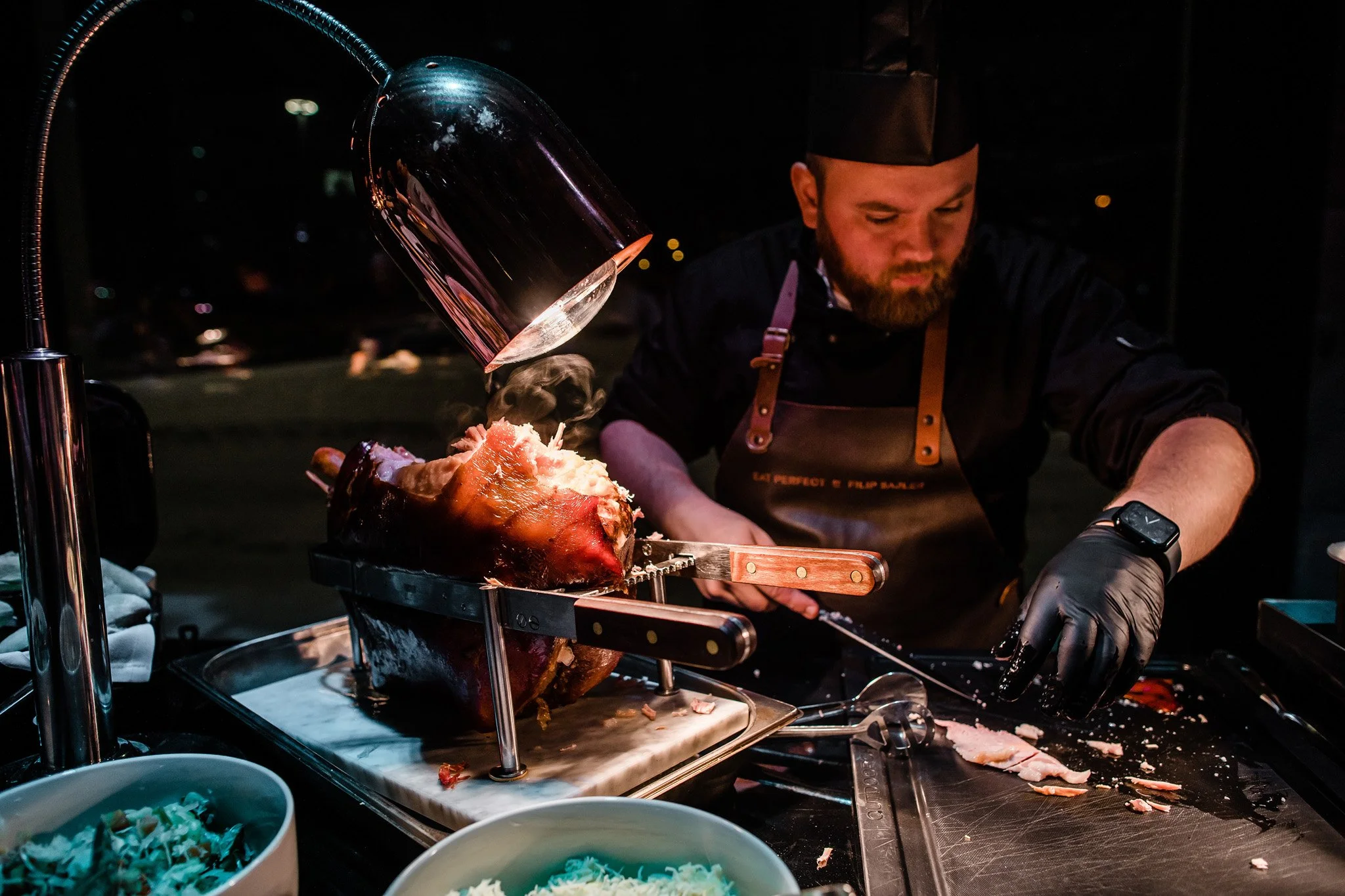 Chef carving a roasted pig at a food event, with a black apron and gloves, in a dark setting. Catering, culinary and food photography in Zürich, Zug, Bern, Lucerne, Basel, St. Gallen and across Switzerland.