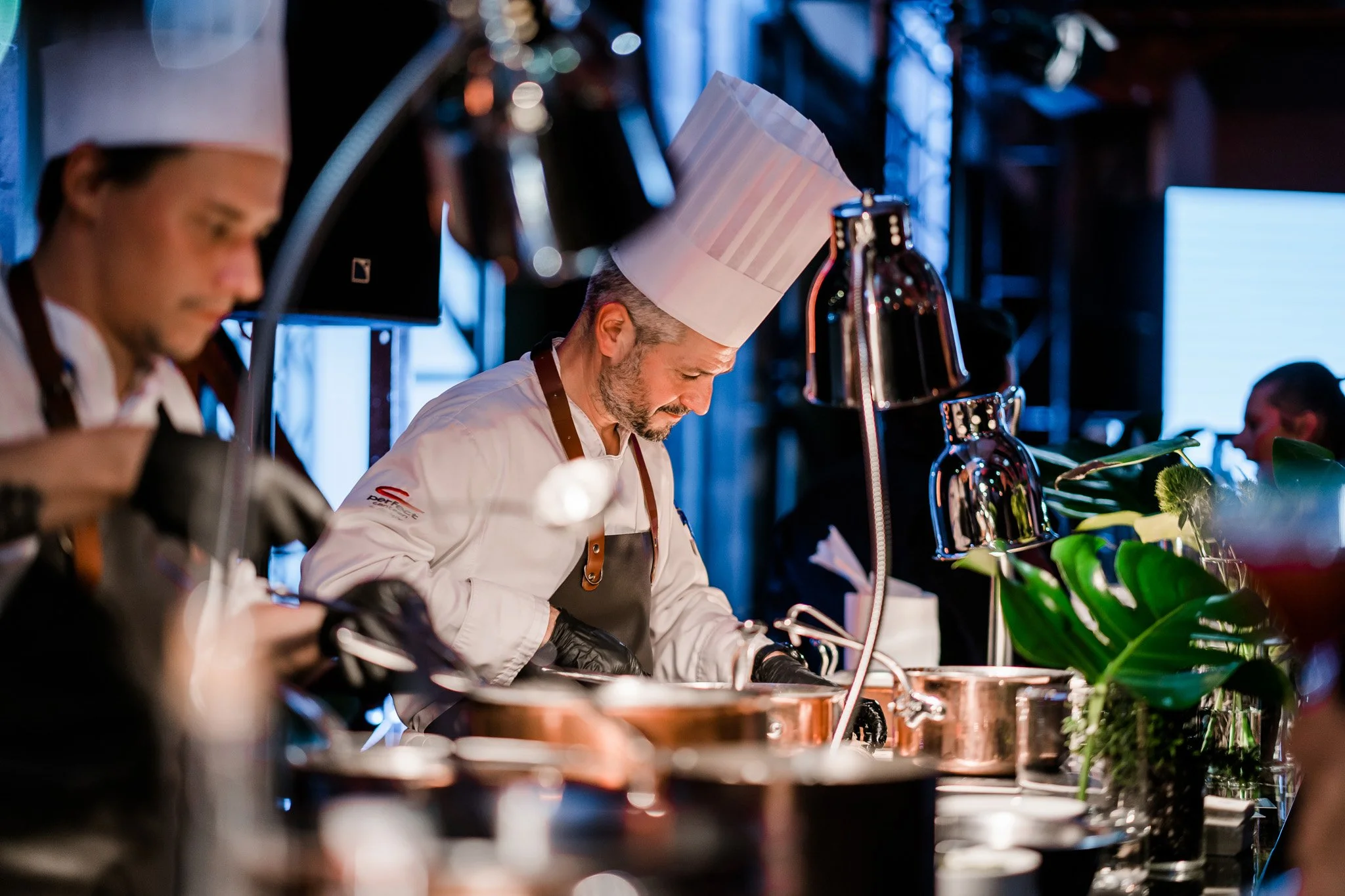 A chef in a tall white hat and white uniform focusing on preparing food in a professional kitchen, with another chef partially visible on the left. Catering and food photography in Zürich, Zug, Bern, Lucerne, Basel, St. Gallen and across Switzerland.