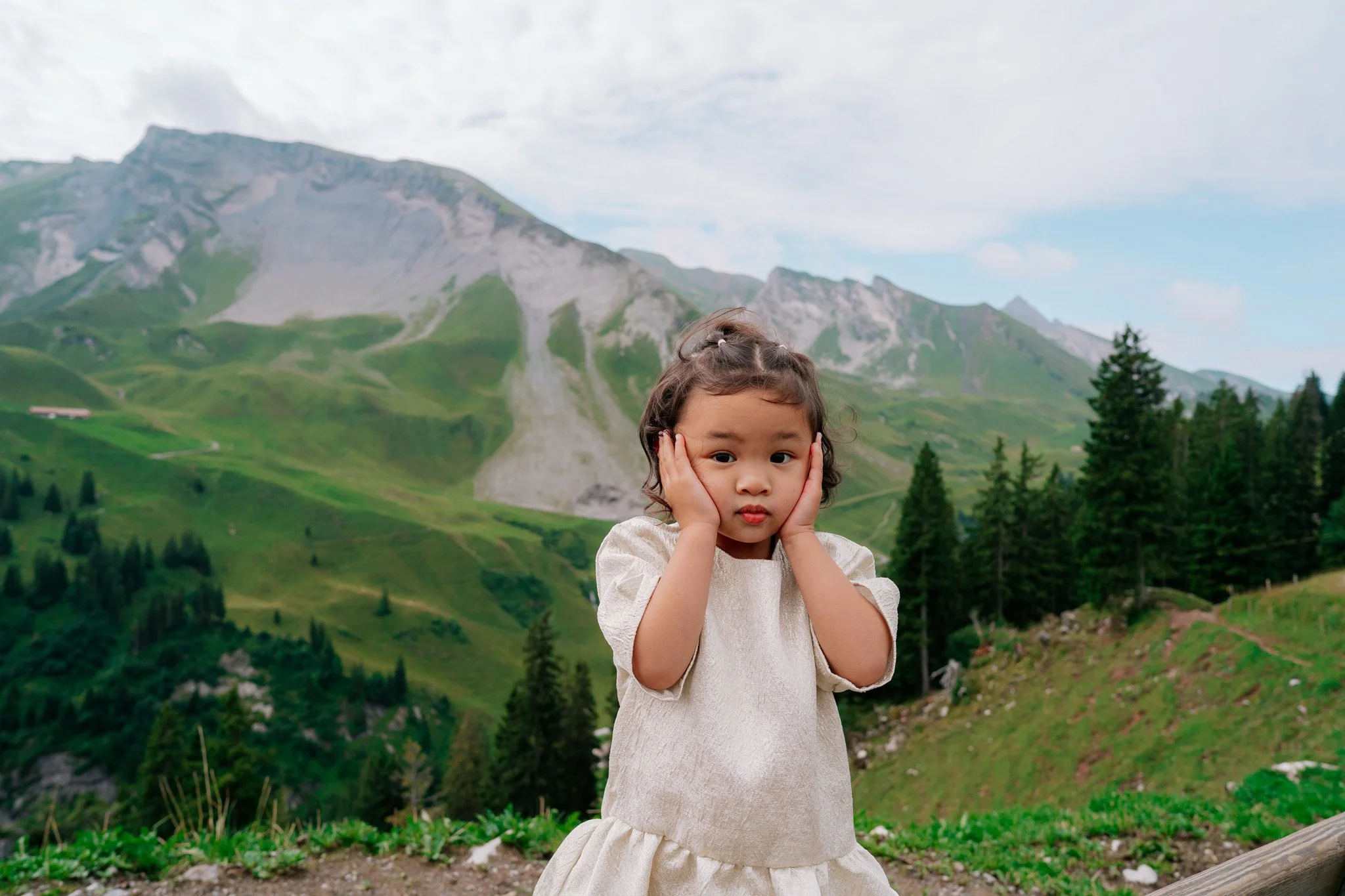 A young girl in a beige dress covering her ears with her hands stands outdoors with a mountainous landscape in the background. Family photoshoot in Switzerland.