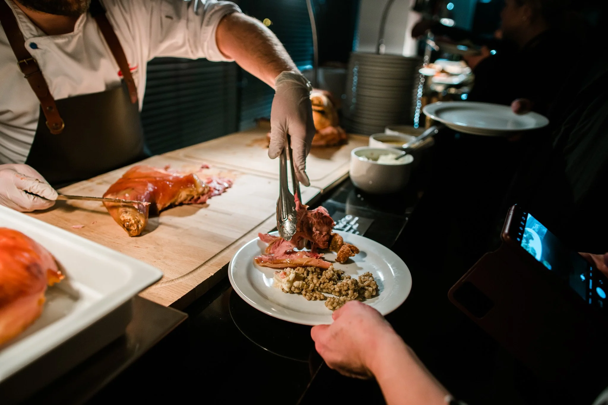 A chef in a white shirt and black apron serves food onto a plate at a buffet-style station with roasted meat and side dishes, while guests wait and take photos. Catering and food photography in Zürich, Zug, Bern, Lucerne, Basel, St. Gallen.