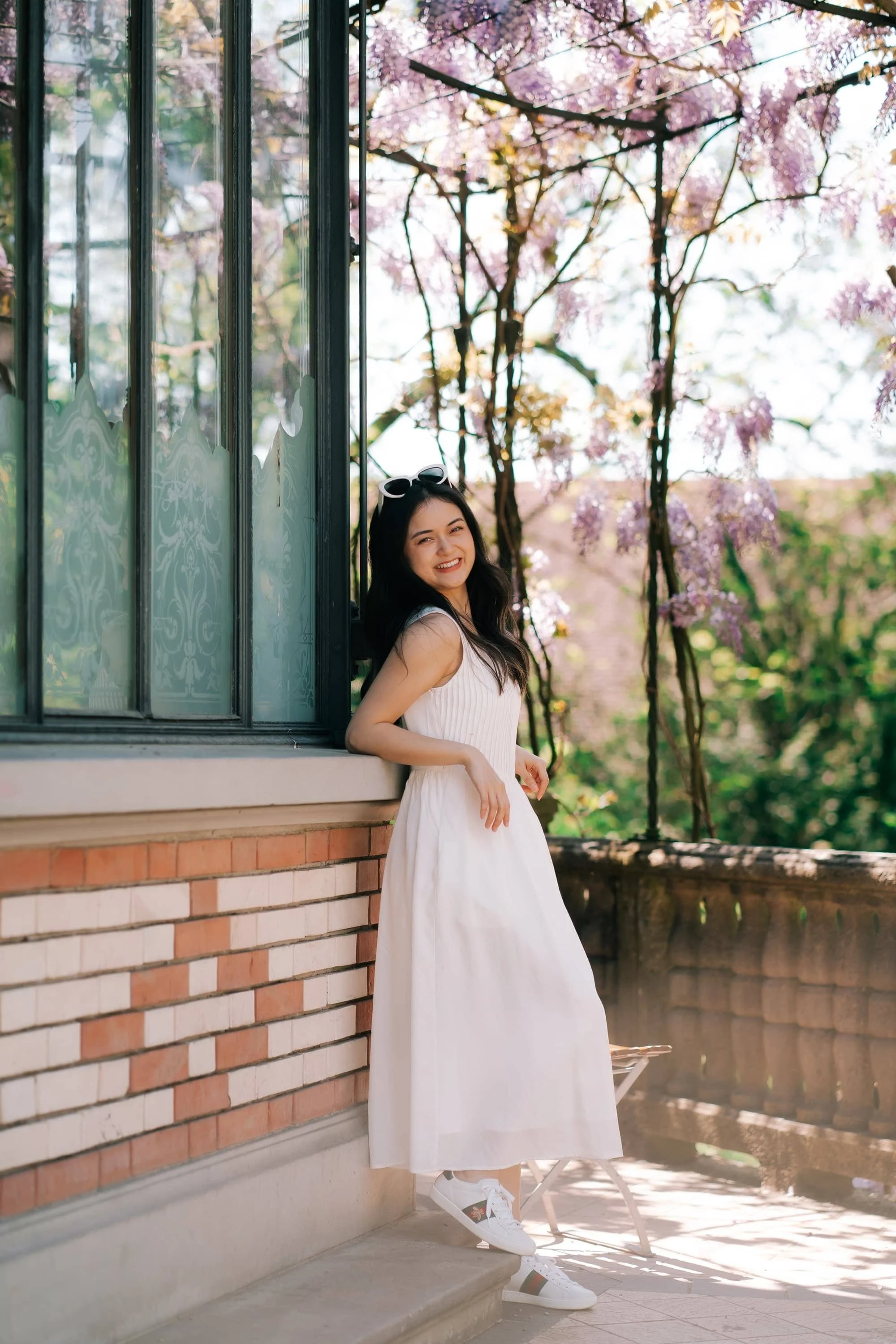 A young woman in a white dress standing outdoors near a window, with flowering trees and a brick wall in the background. Villa Patumbah gardens in Zurich.