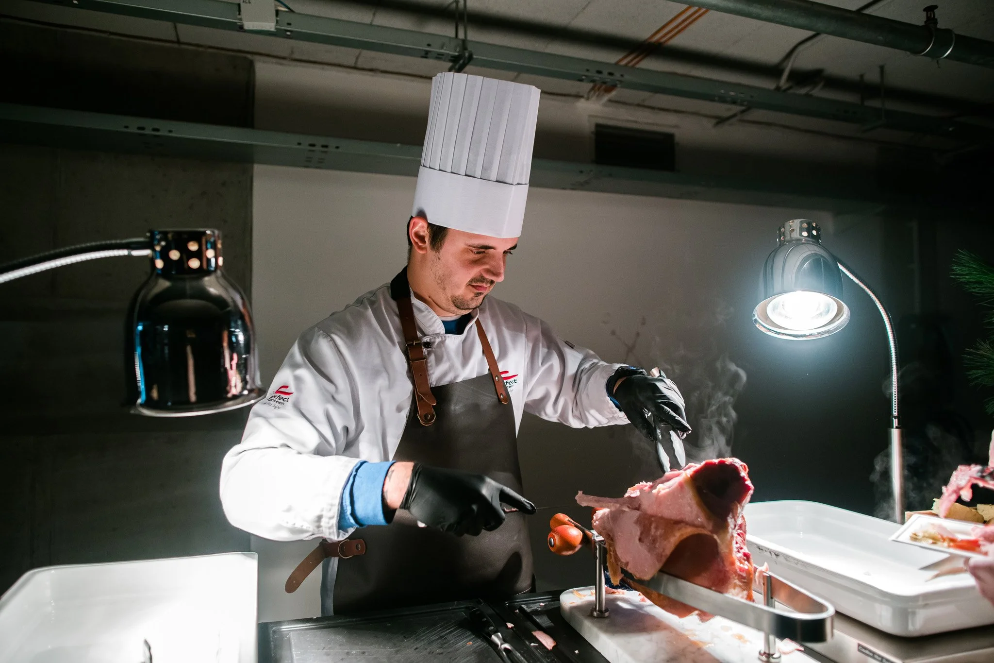 Chef in a tall white hat and apron carving a large piece of meat in a professional kitchen. Catering and food photography in Zürich, Zug, Bern, Lucerne, Basel, St. Gallen and across Switzerland.