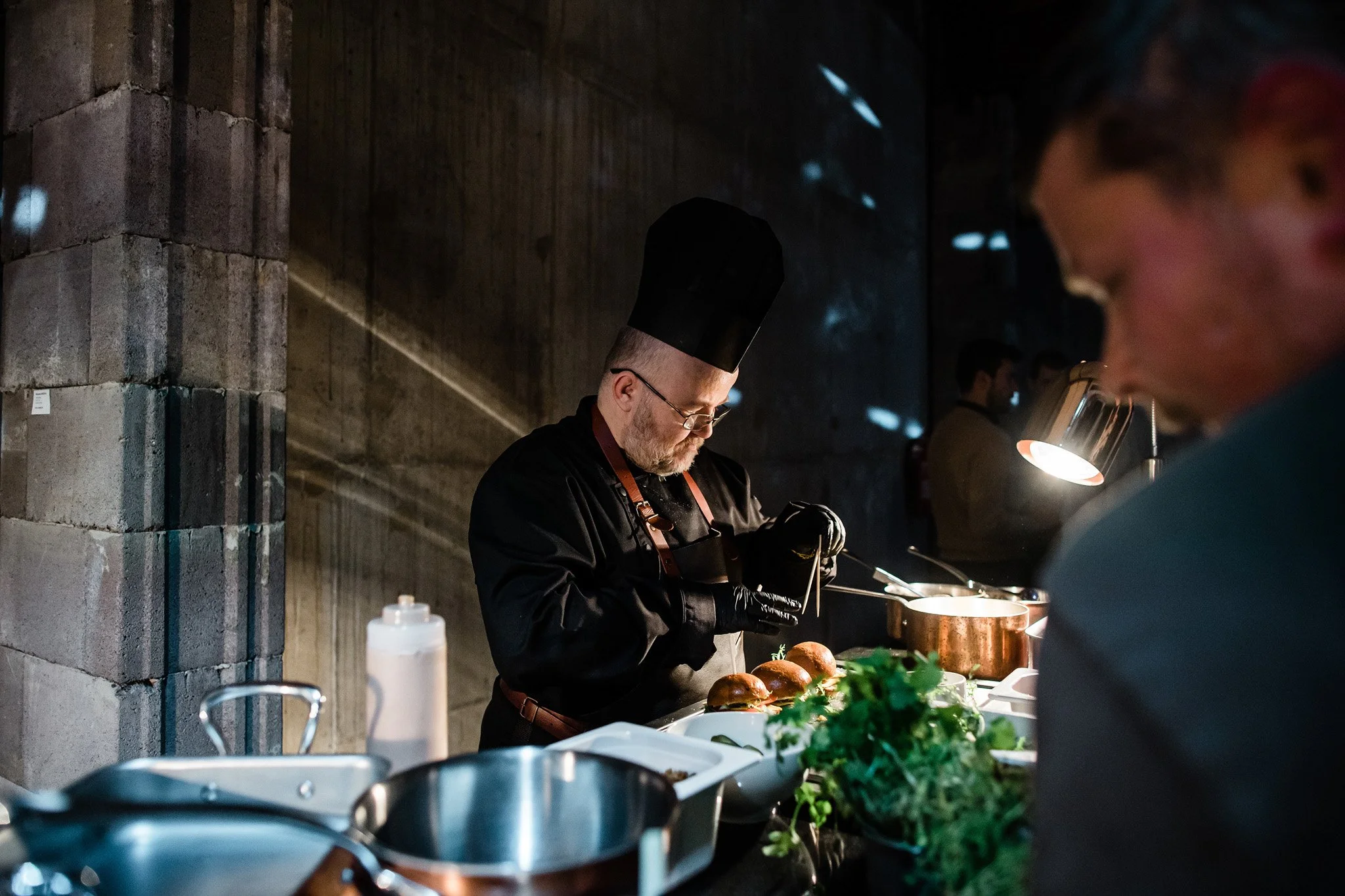 A chef in black uniform and tall black hat preparing food at a professional kitchen station, with a customer in the foreground. Catering and food photography in Zürich, Zug, Bern, Lucerne, Basel, St. Gallen and across Switzerland.