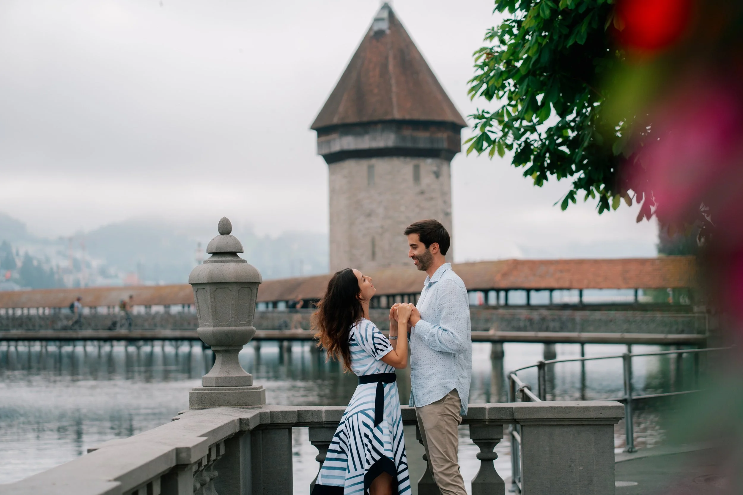 A couple holding hands and smiling at each other by a river in Lucerne Switzerland with an old stone tower and bridge in the background.