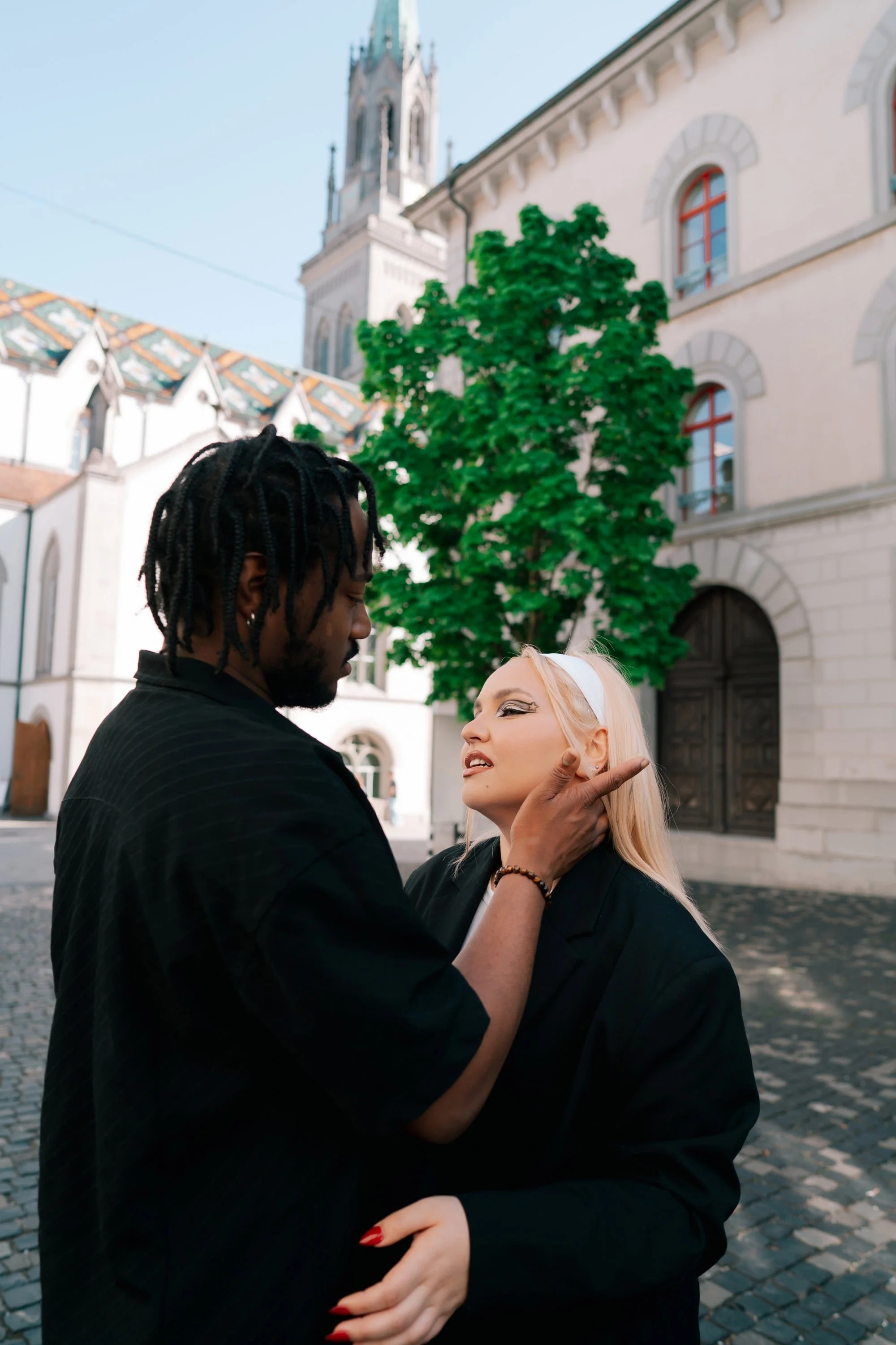 A man with dreadlocks holds the chin of a blonde woman with makeup and a white headband, outside in front of a historic European building in St Gallen Switzerland with arched windows and a tower, on a cobblestone street.