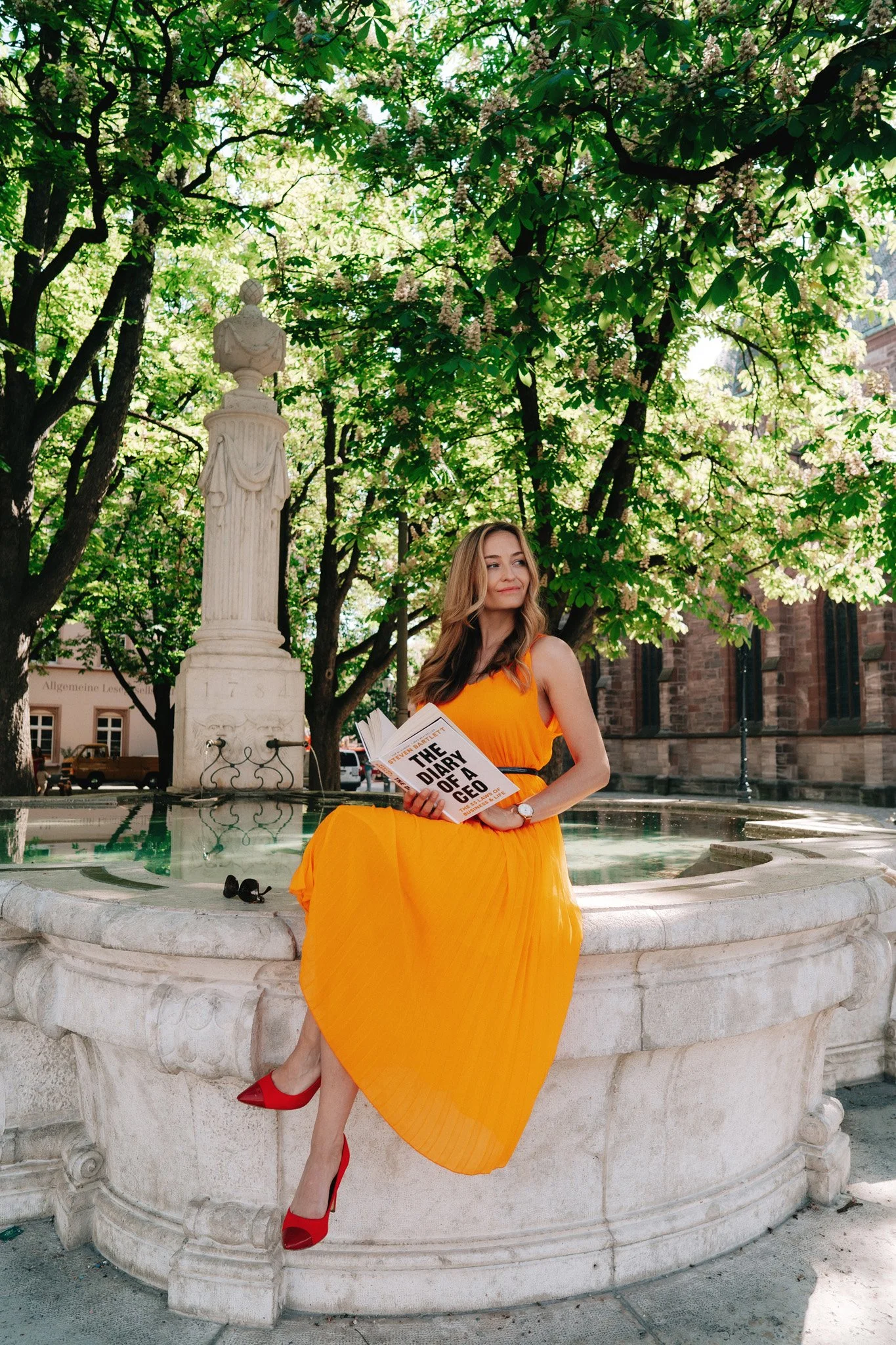Solo portrait photoshoot of woman in a flowing orange dress and red heels sitting on the edge of a stone fountain in a park in Basel, Switzerland, holding a book titled 'The Diary of a CEO', with green trees and a classical statue in the background.